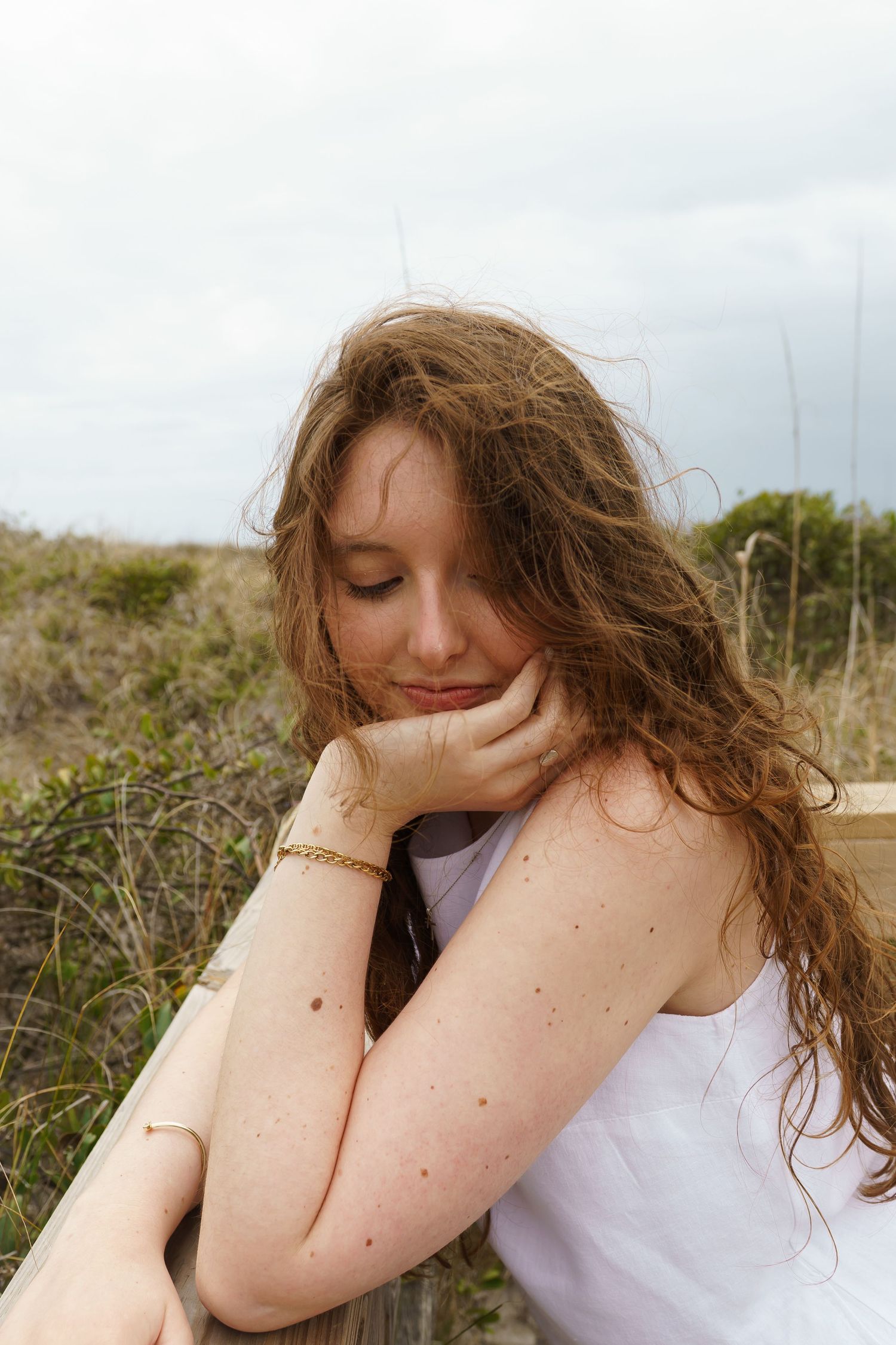 Young woman with long wavy hair resting chin on hand, leaning on wooden railing outdoors near coastal dunes.