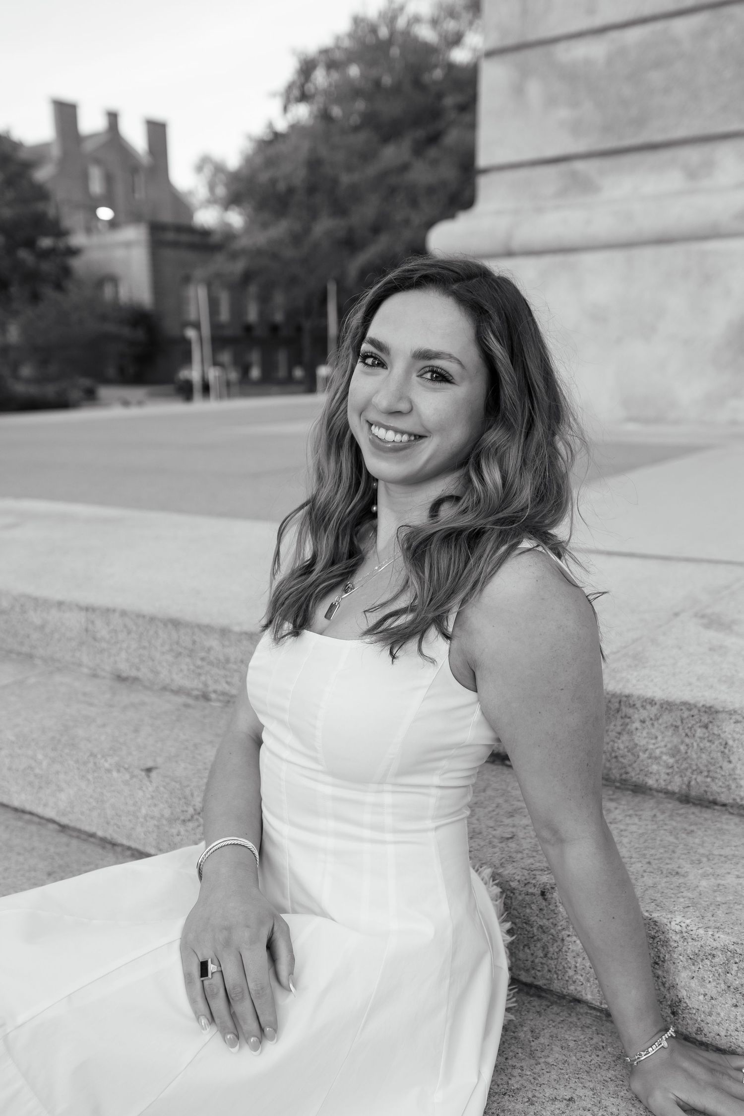 Black and white photo of a smiling young woman in a white dress sitting on stone steps outdoors.