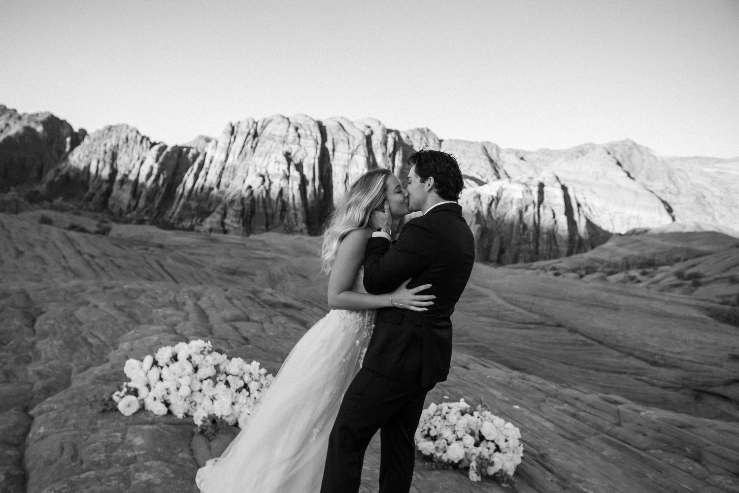 Wedding couple embraces among white floral arrangements with dramatic red rock cliffs in background.
