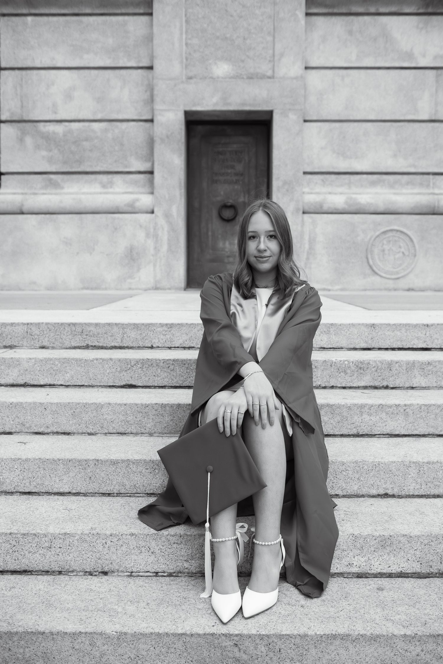 Black and white photo of a young woman in graduation gown and heels sitting on stone steps holding her cap.
