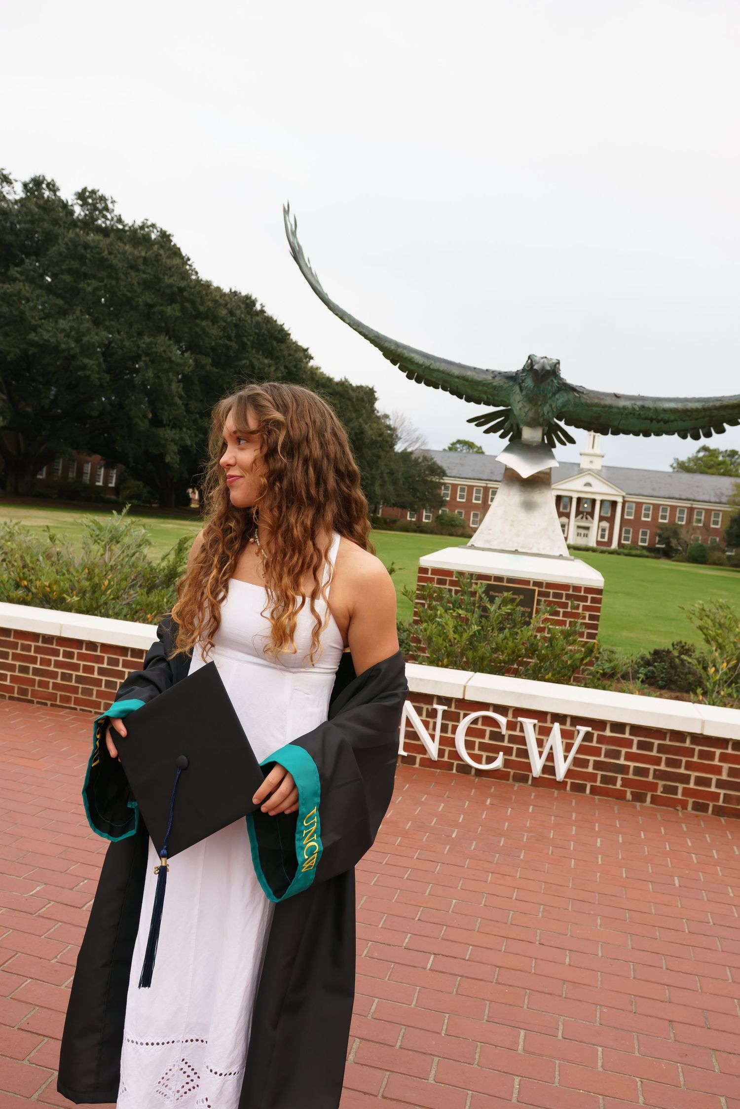 A graduate in a white dress and black gown poses with a hawk statue at UNCW campus.
