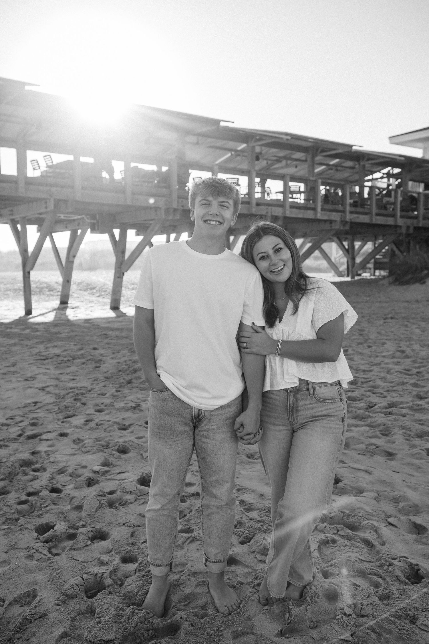 Black and white photo of couple standing on beach near wooden pier.