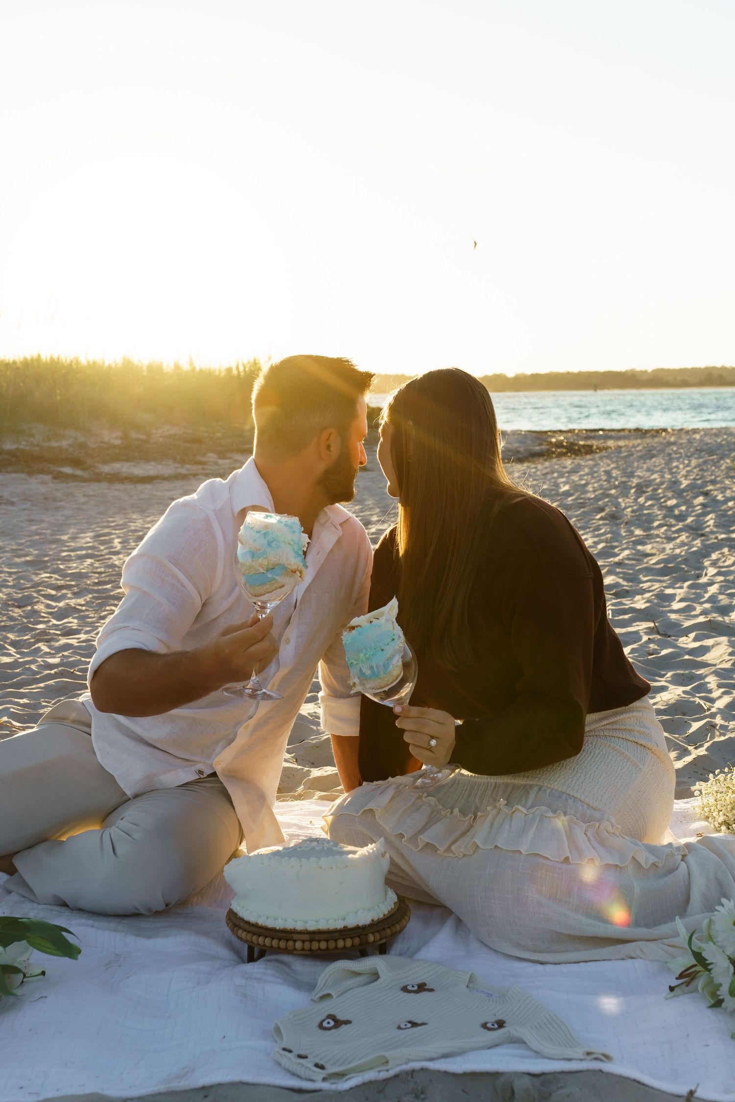 Romantic sunset beach picnic with a couple sharing drinks and cake on a white blanket.