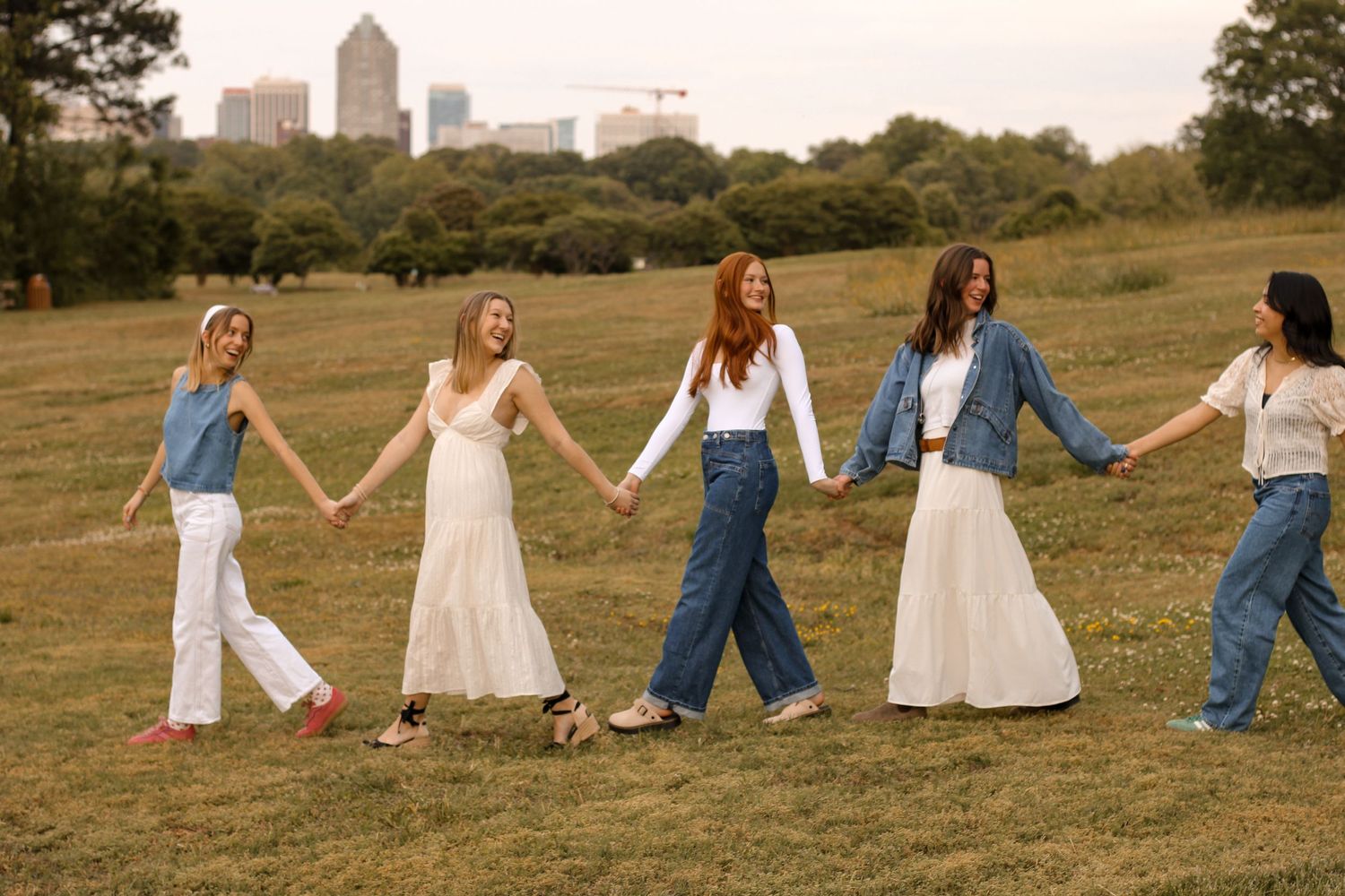 Five friends holding hands walk in a line across grassy field with urban skyline behind them.
