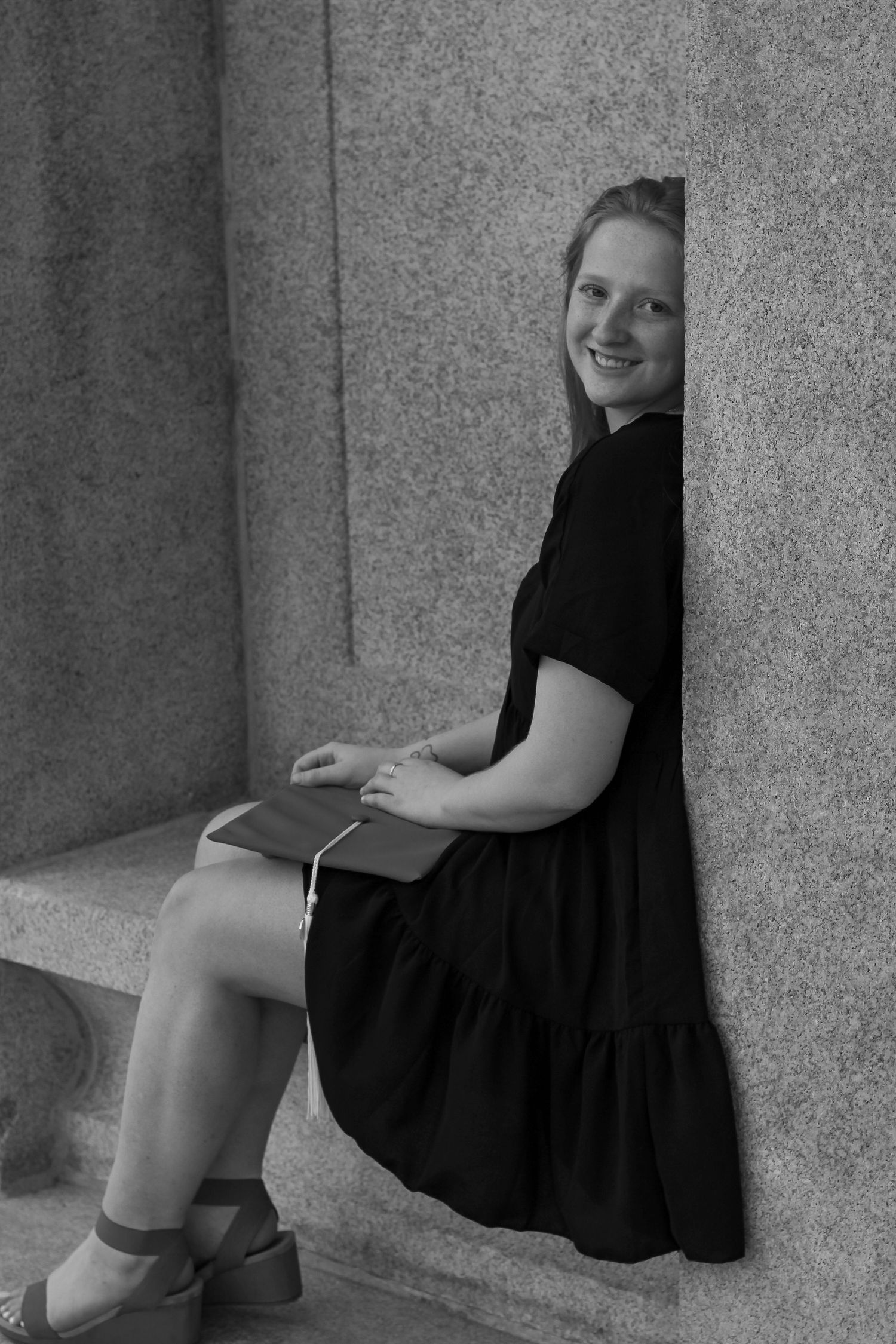 Black and white photo of graduate in casual dress seated on stone ledge.