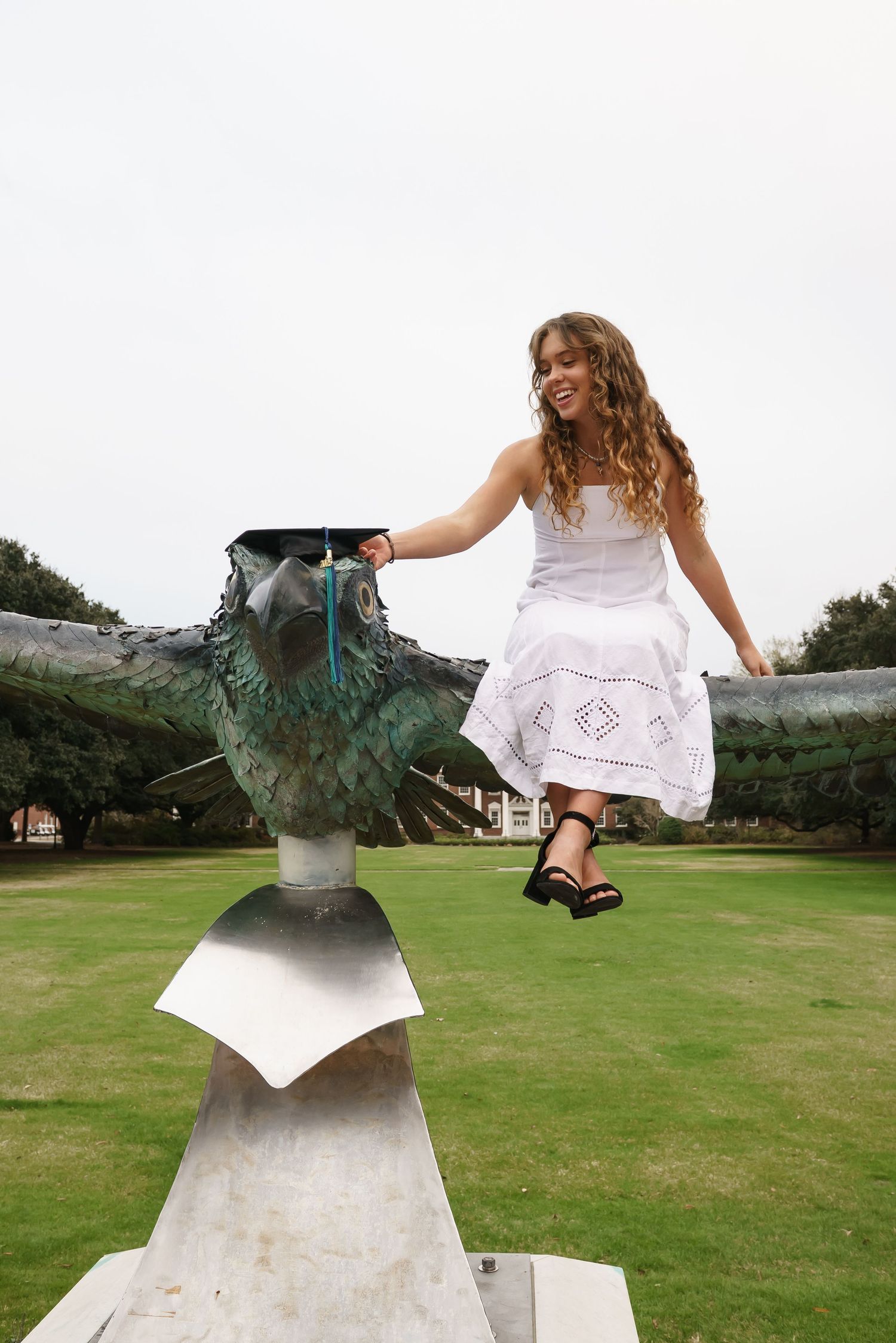 Graduate poses playfully on campus sculpture wearing white dress and black heels.