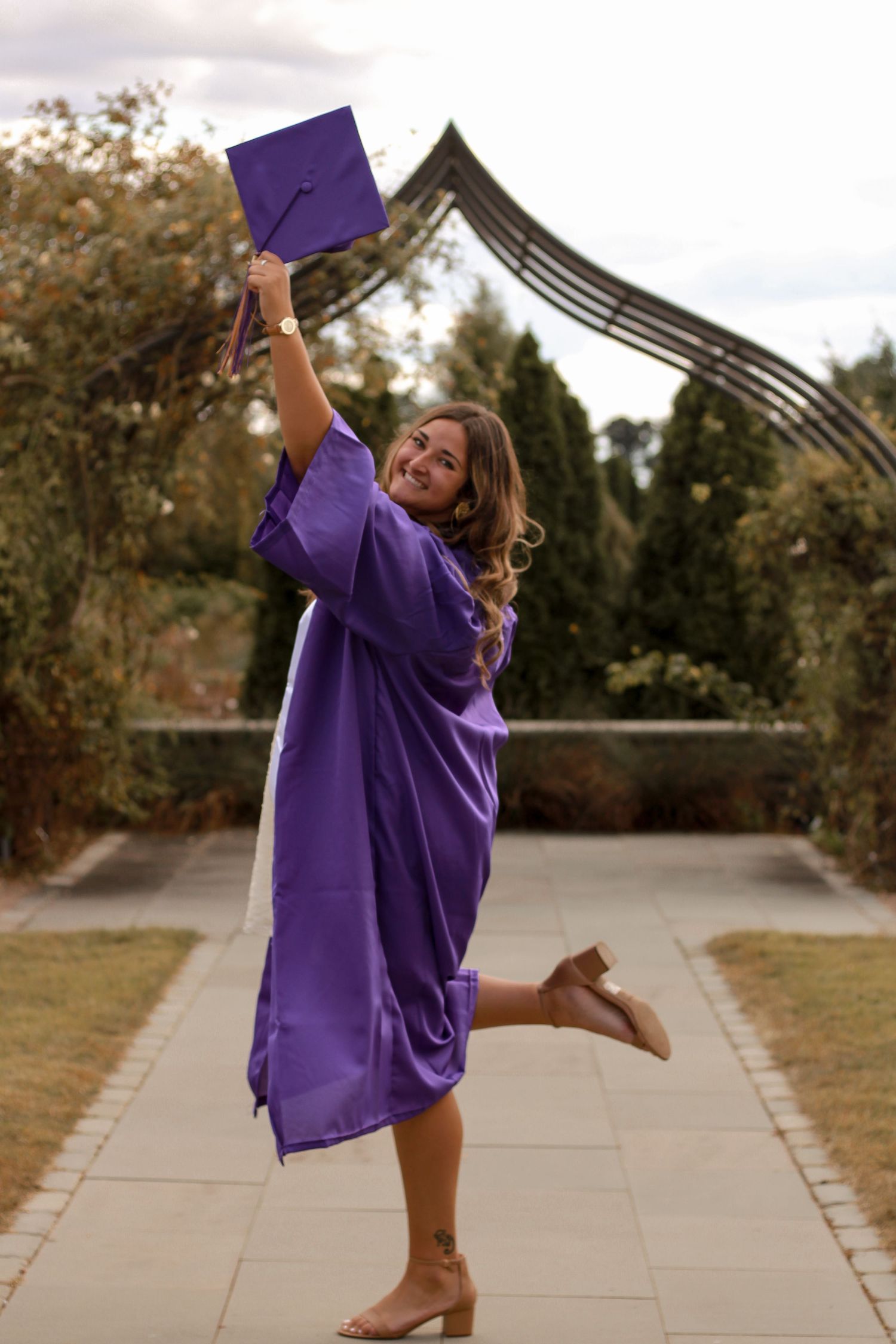 Graduate in purple cap and gown celebrates by jumping and raising diploma against architectural backdrop.
