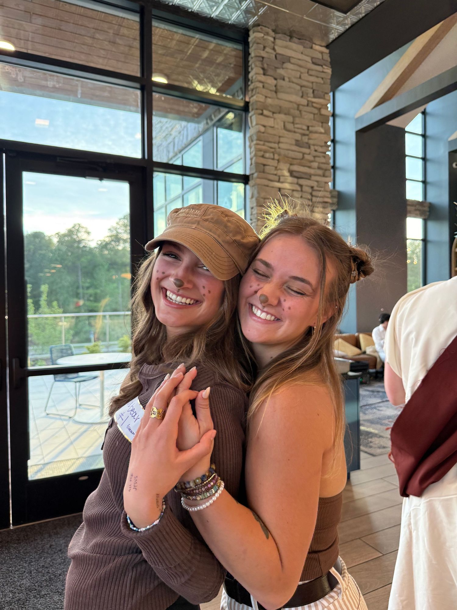 Two young women smiling and hugging indoors, one wearing a brown cap, near large windows with a wooded view.
