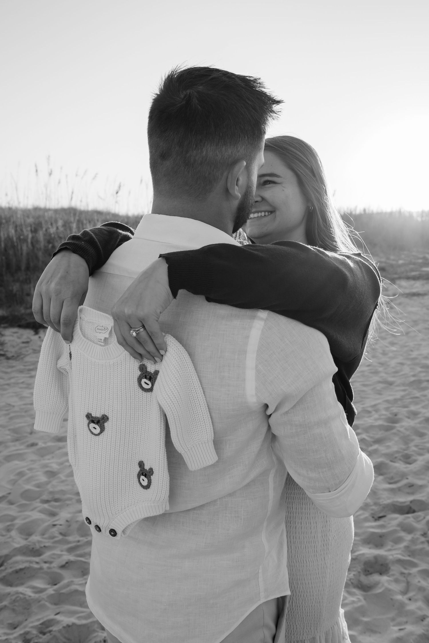 Black and white photo of a joyful embrace on the beach with a baby onesie.