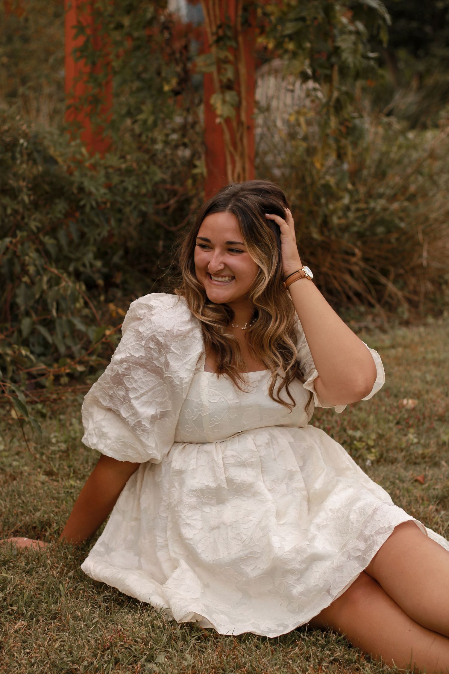 Person in white eyelet dress sits on grass while smiling and touching hair.
