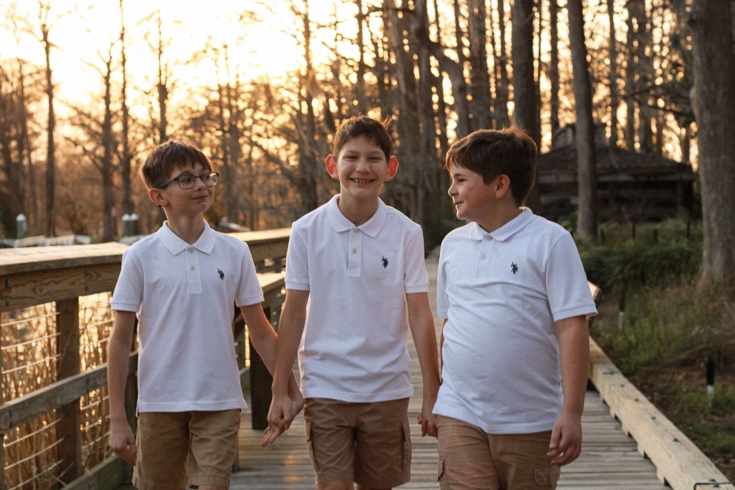 Three boys in matching white polo shirts walk together on a wooden boardwalk at sunset.
