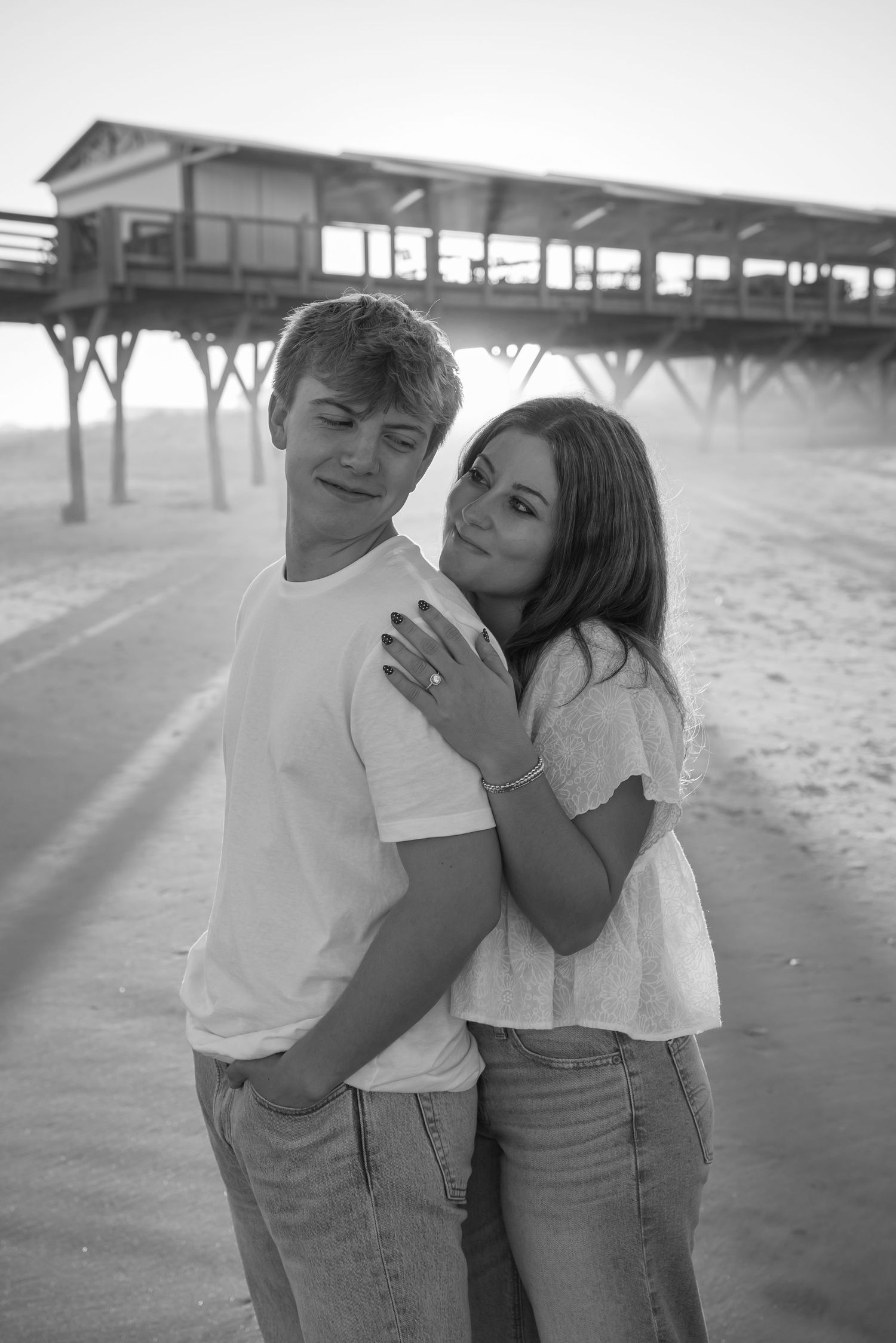 A black and white portrait of a couple embracing on the beach with a pier in the background.
