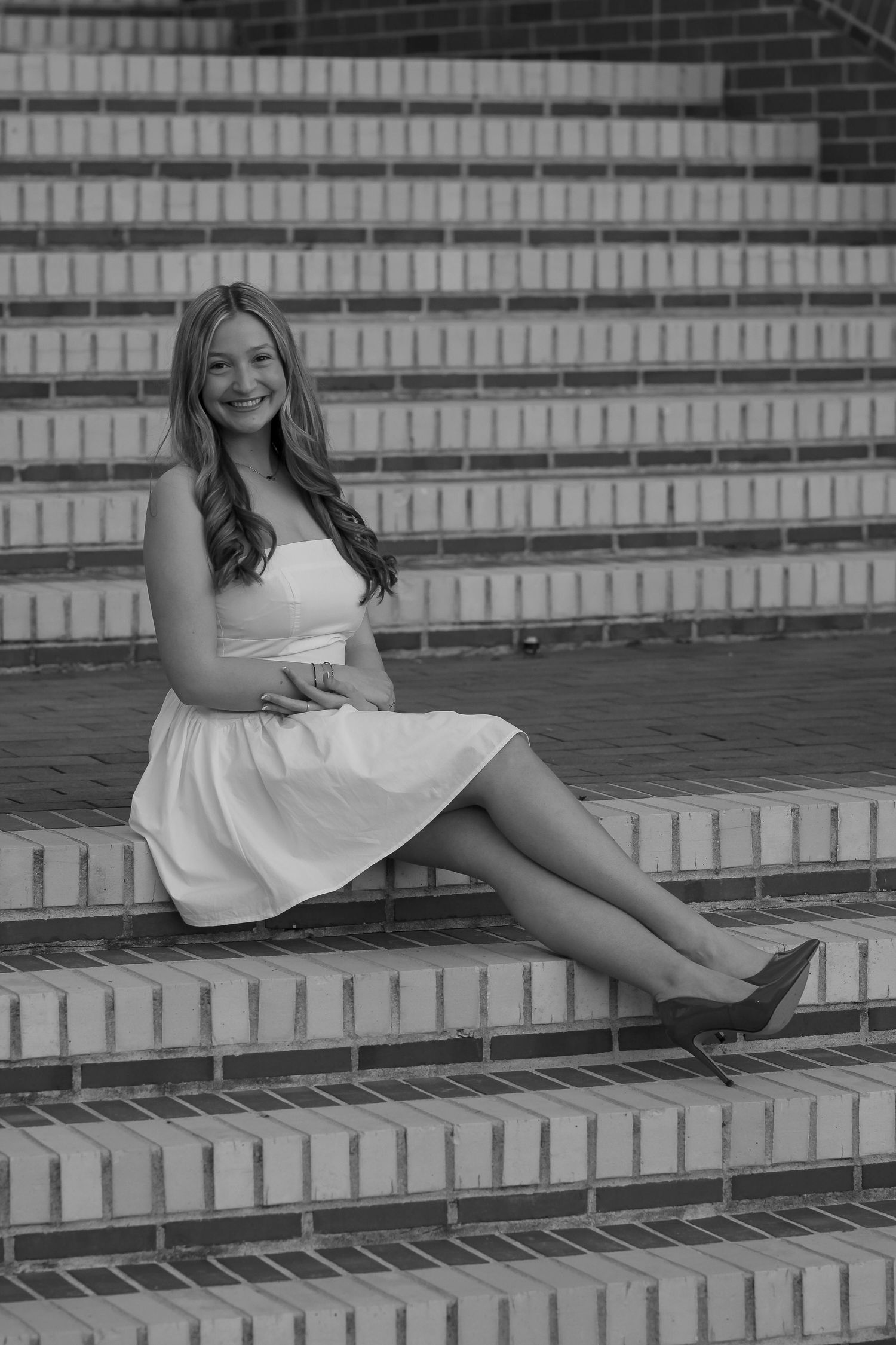 Graduate in white dress sitting on brick steps in black and white photography.