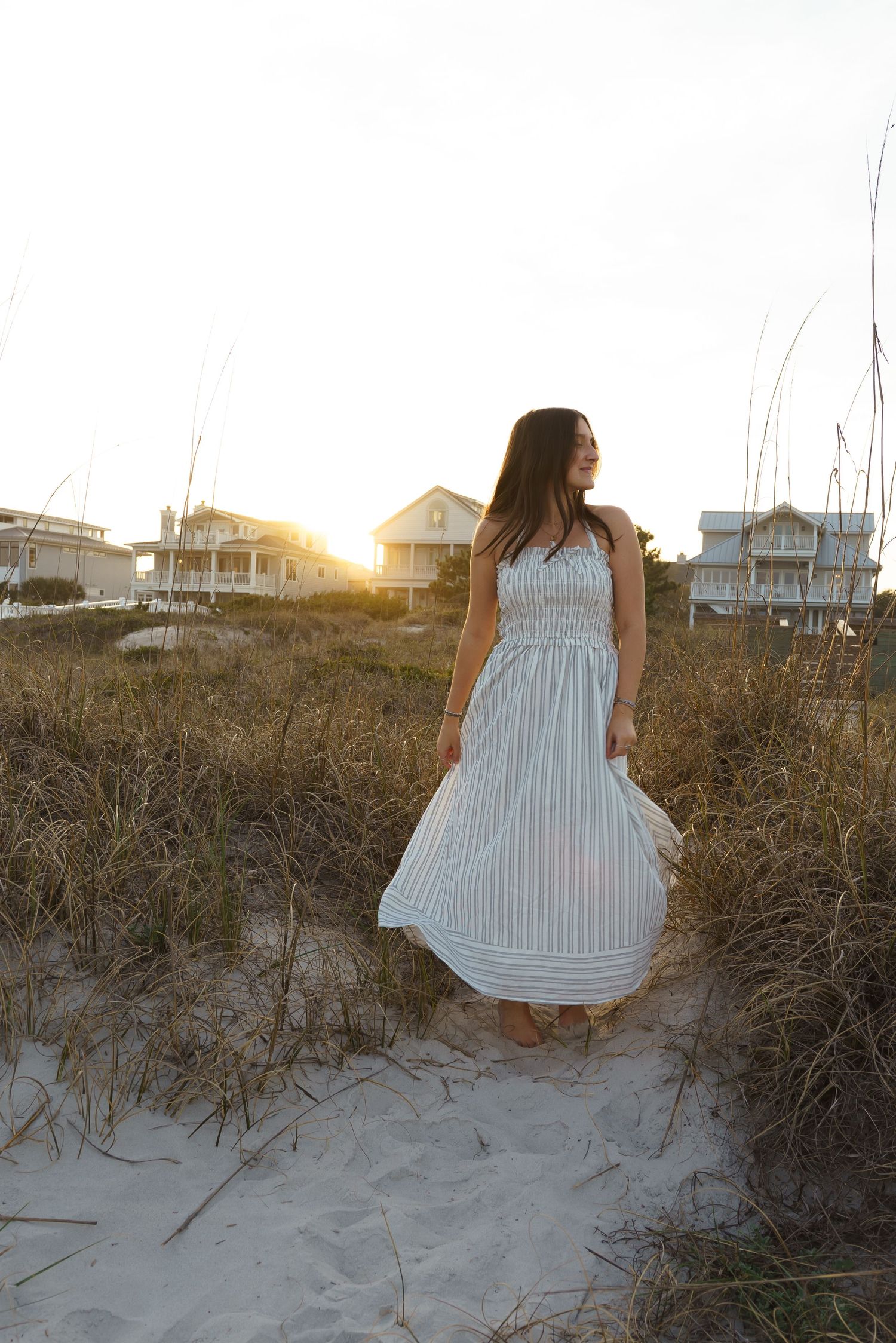 Woman in striped maxi dress standing on sandy beach path at golden sunset with coastal houses in background.