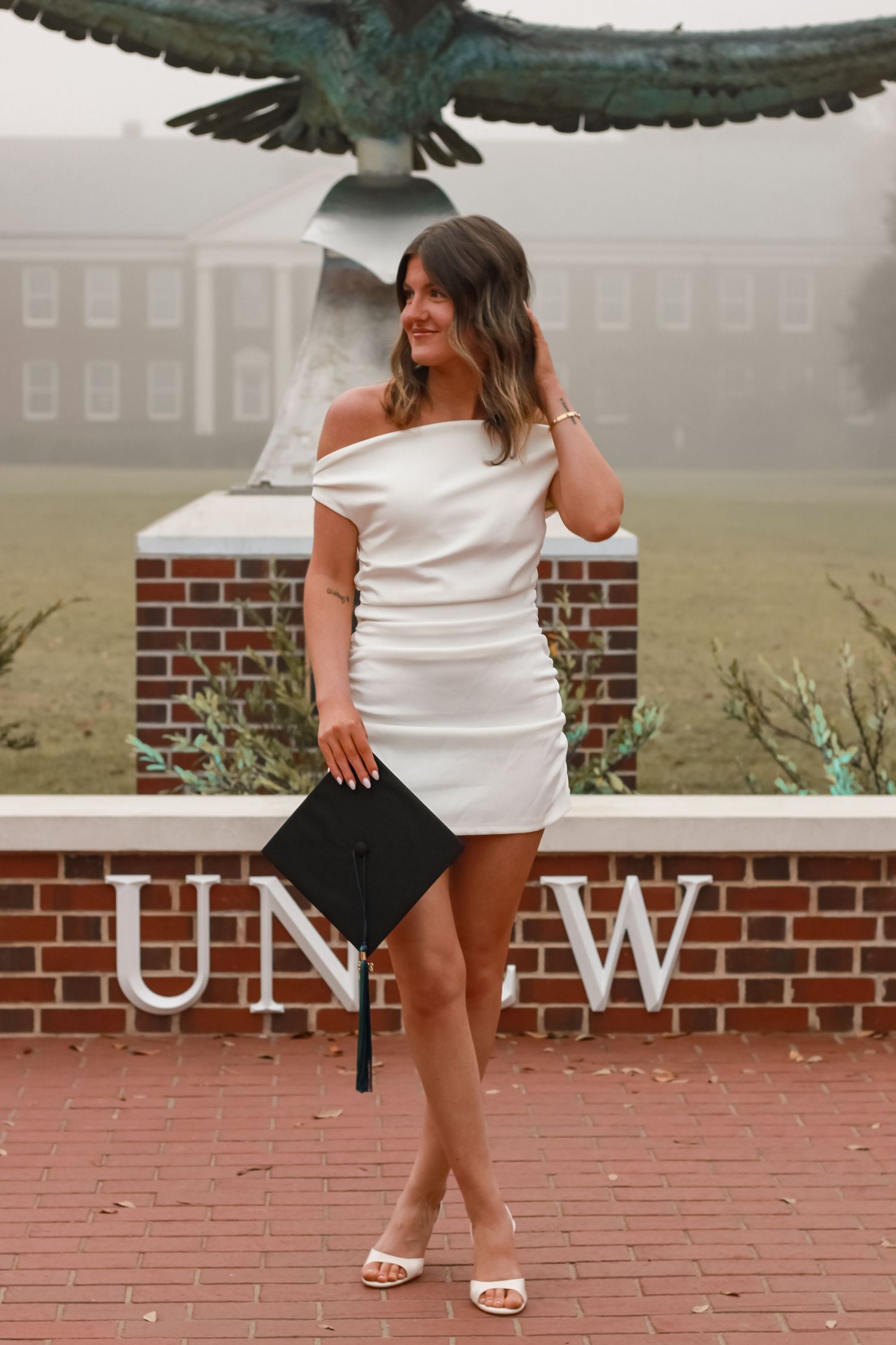 Graduate in white dress posing with diploma by UNCW eagle monument.