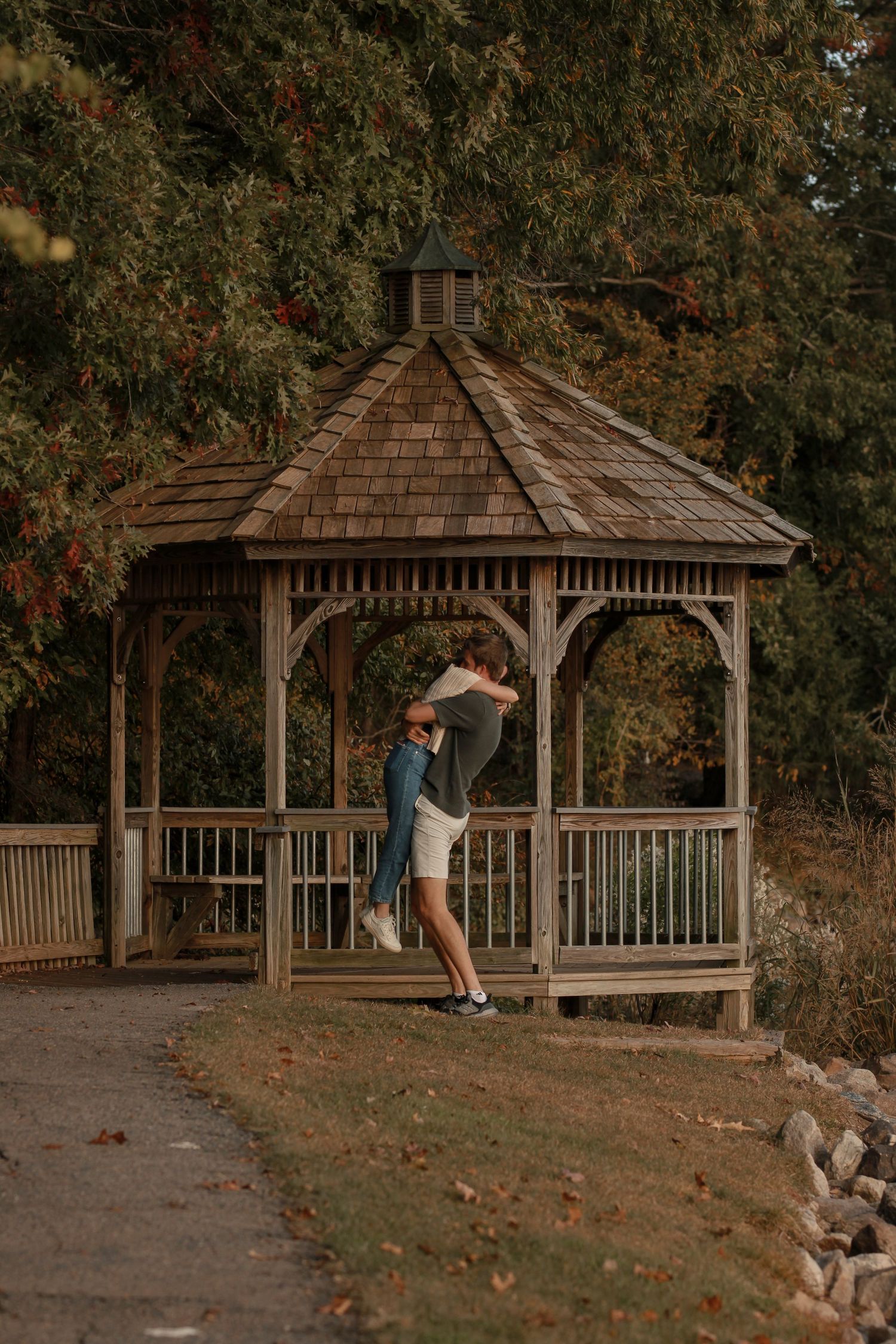 A romantic couple shares a tender moment in front of a rustic gazebo surrounded by autumn trees.