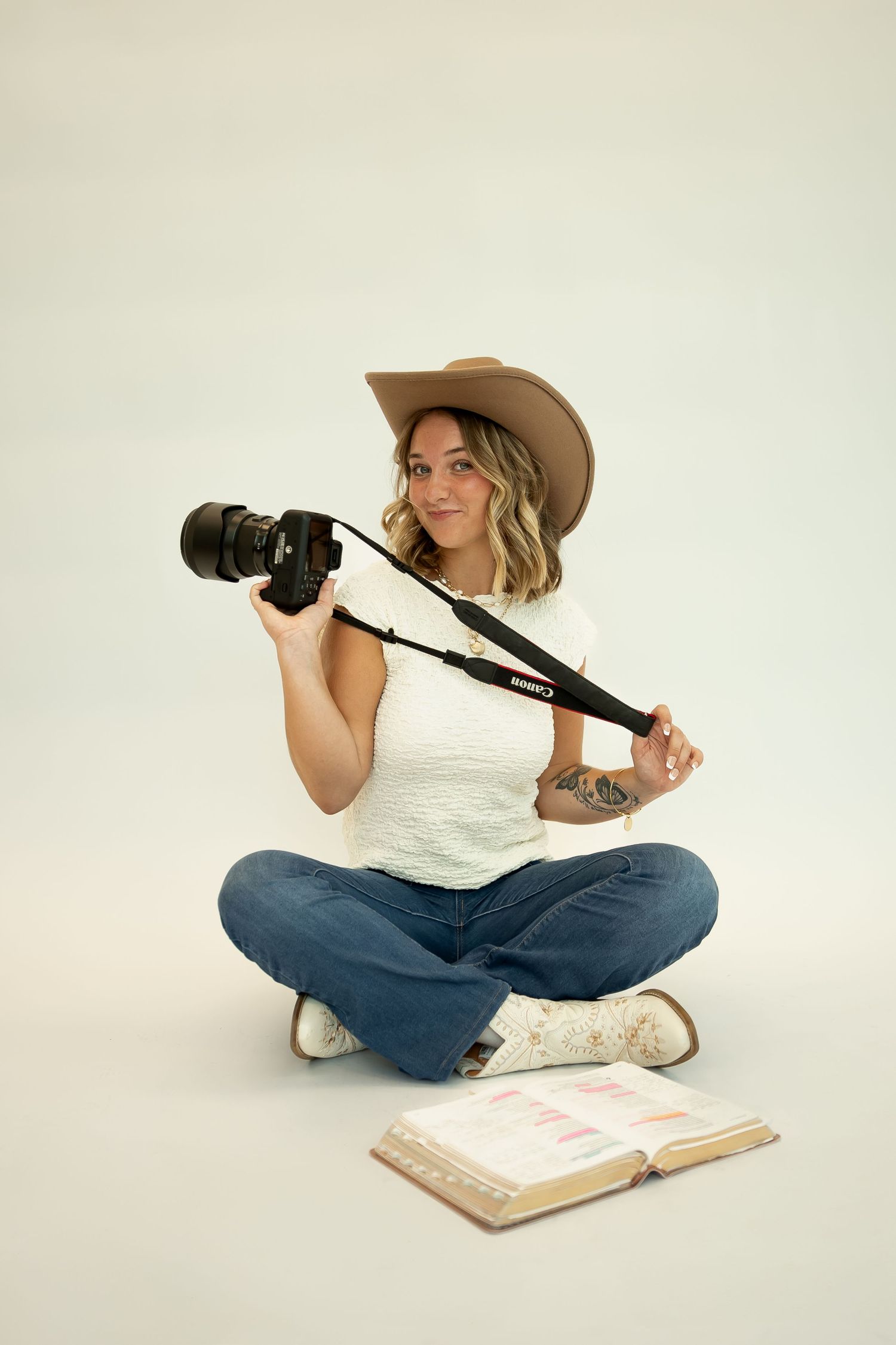 A photographer in casual attire sits cross-legged with camera equipment and books on white background.