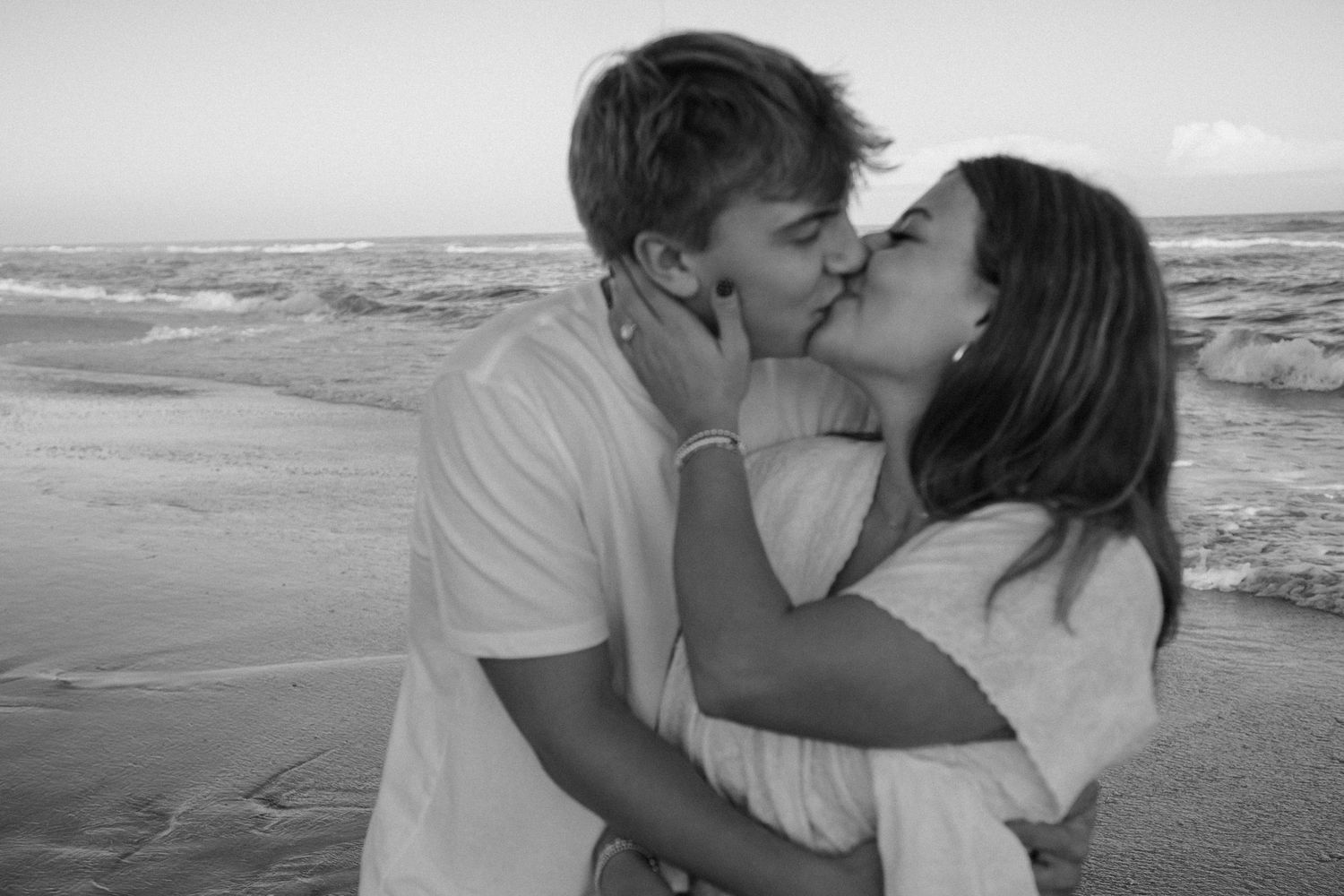 A black and white photo of a couple sharing a romantic kiss on the beach.