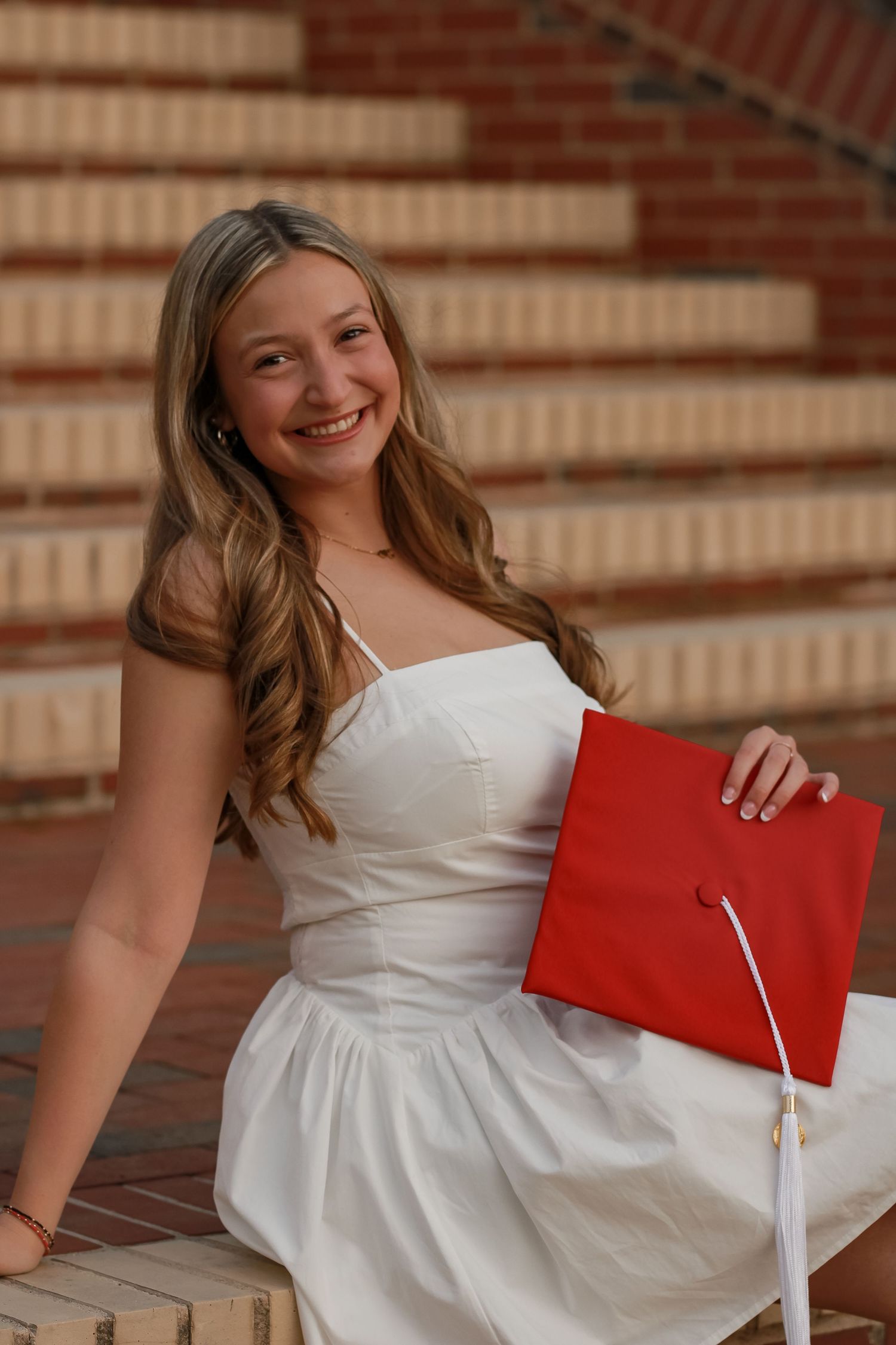 Graduate in white dress holding red diploma cover while sitting on brick steps.