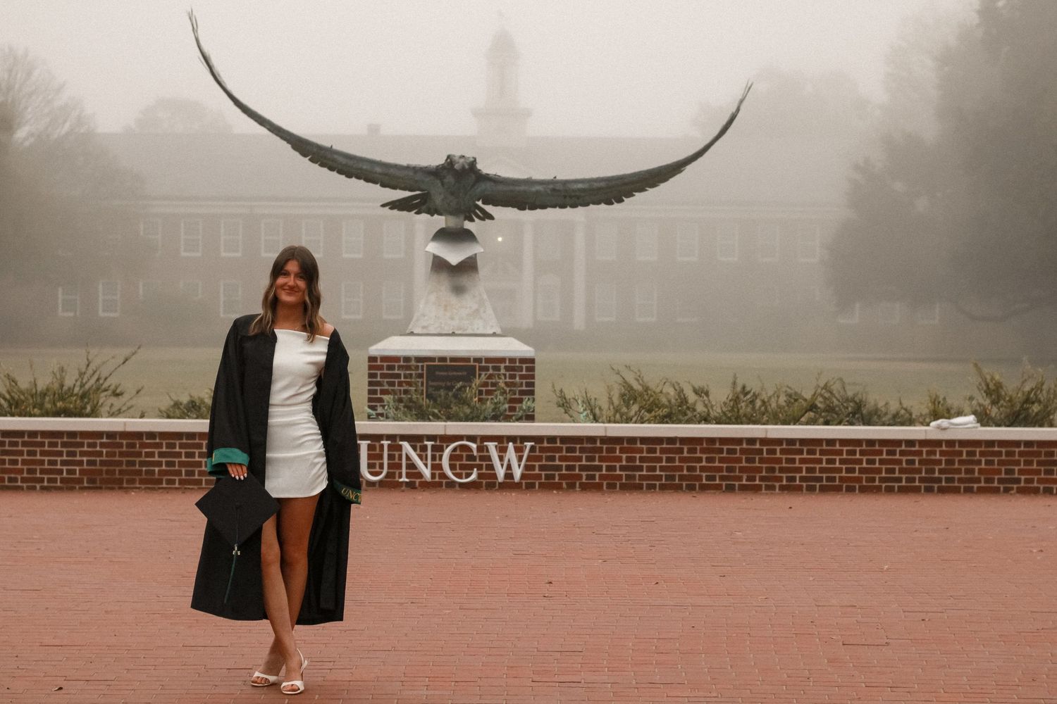 Graduate in graduation gown standing by UNCW eagle statue on misty morning.