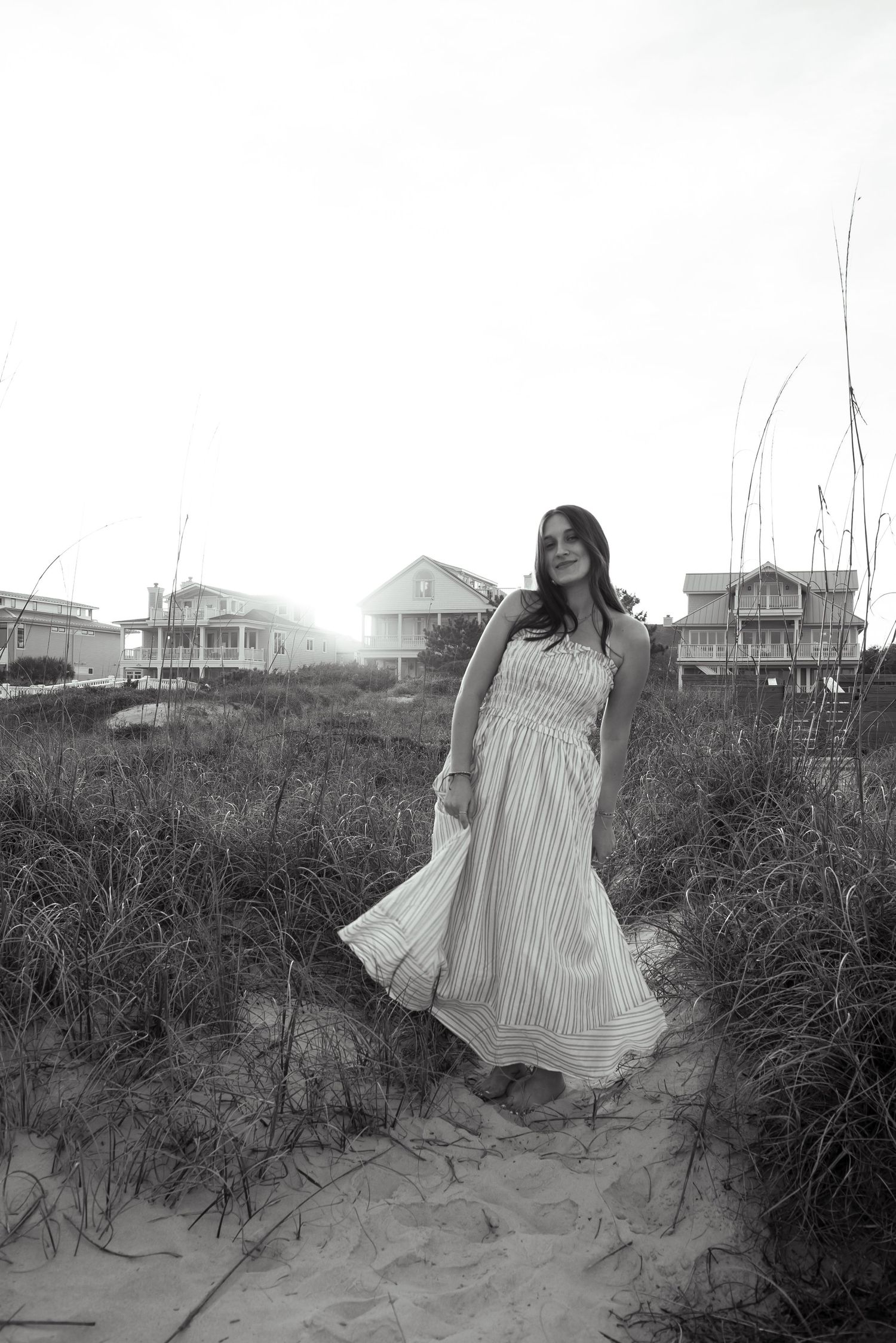 Black and white photo of a woman in a flowing striped maxi dress standing on a sandy beach path with coastal homes behind her.