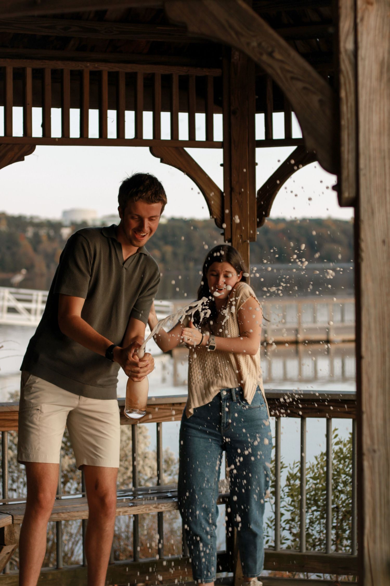 A playful couple splashes water at each other under a wooden gazebo by the waterfront at sunset.