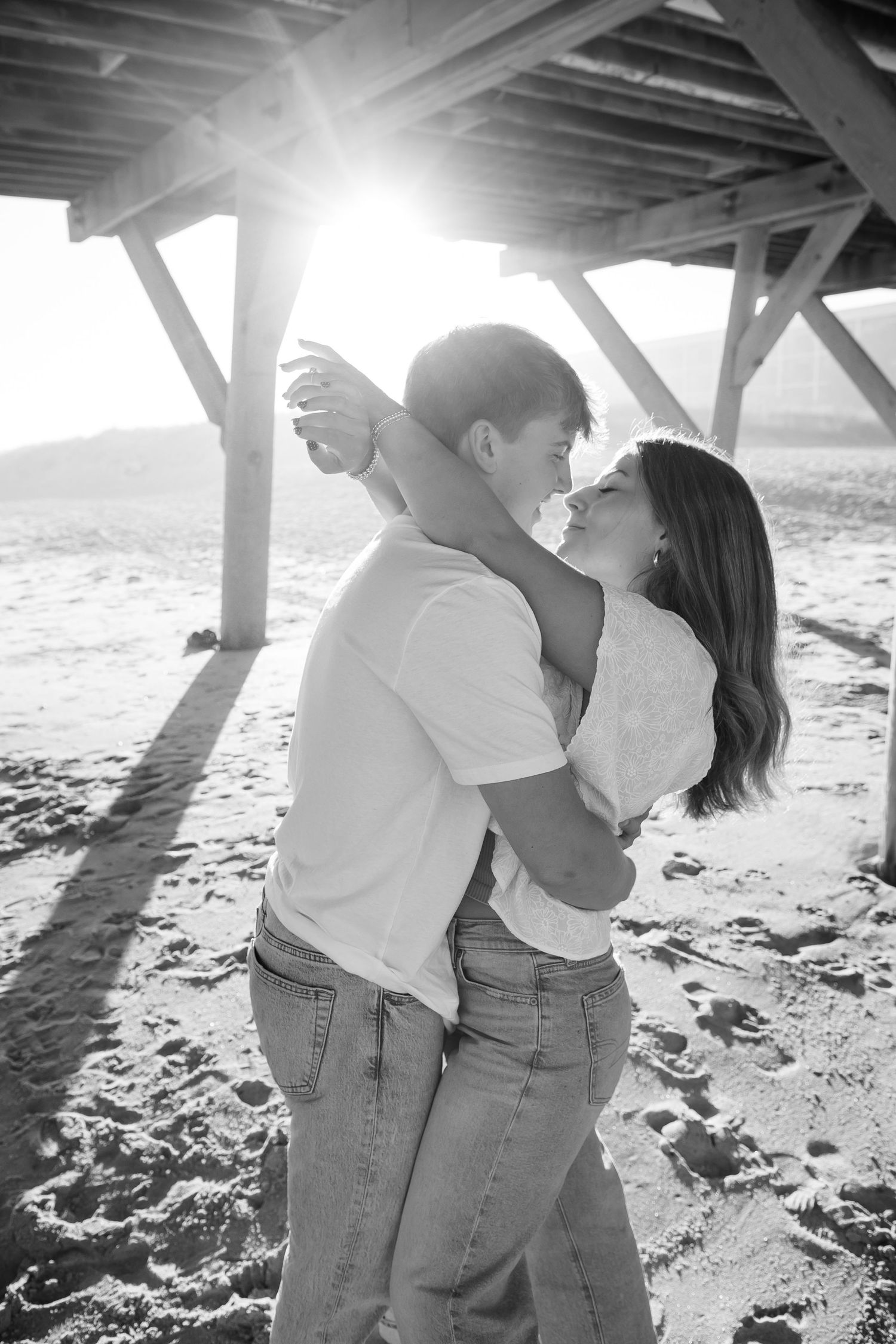 Couple sharing intimate moments under wooden pier supports in black and white photography.