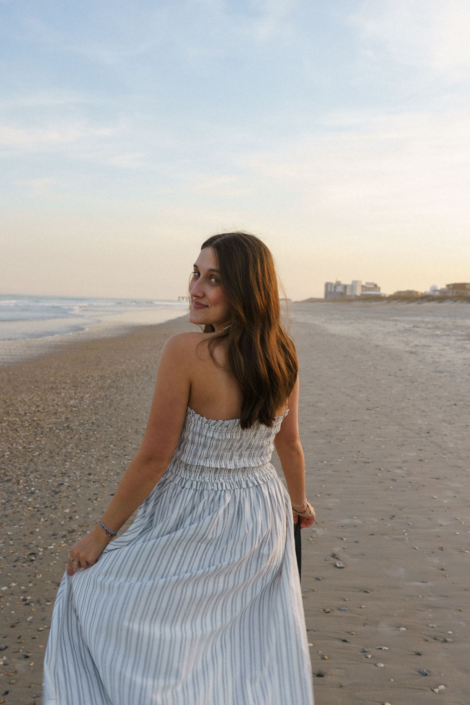 Young woman in striped sundress walks along sandy beach at sunset, glancing back over her shoulder.