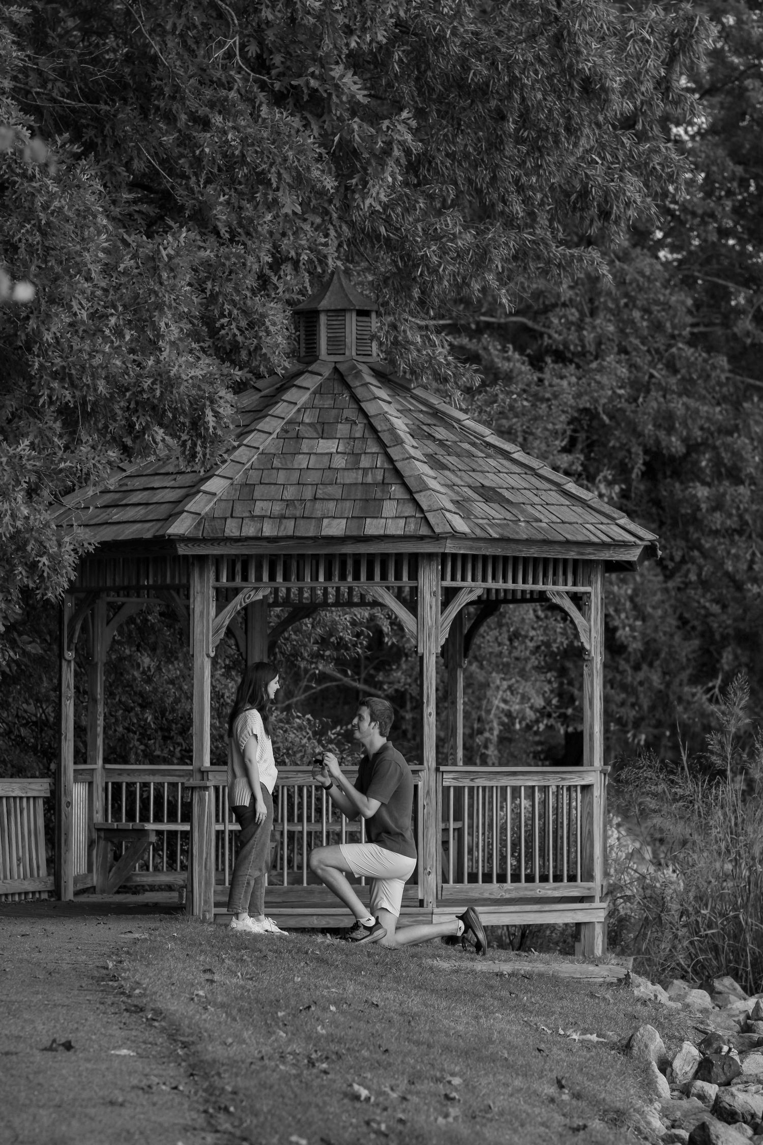 Black and white photo of proposal moment in wooden gazebo surrounded by trees.