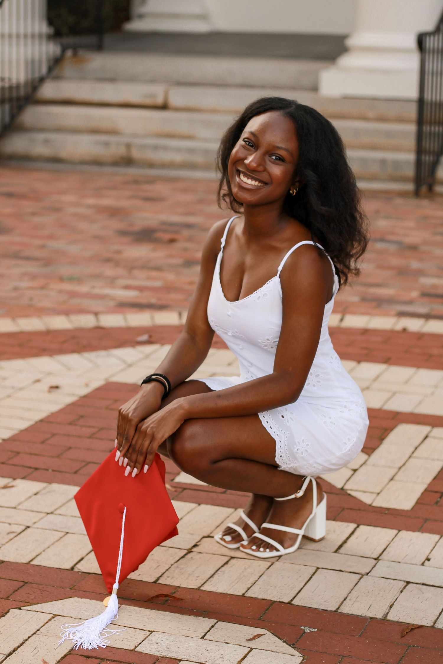 Graduate in white dress poses with red diploma cover on brick courtyard.