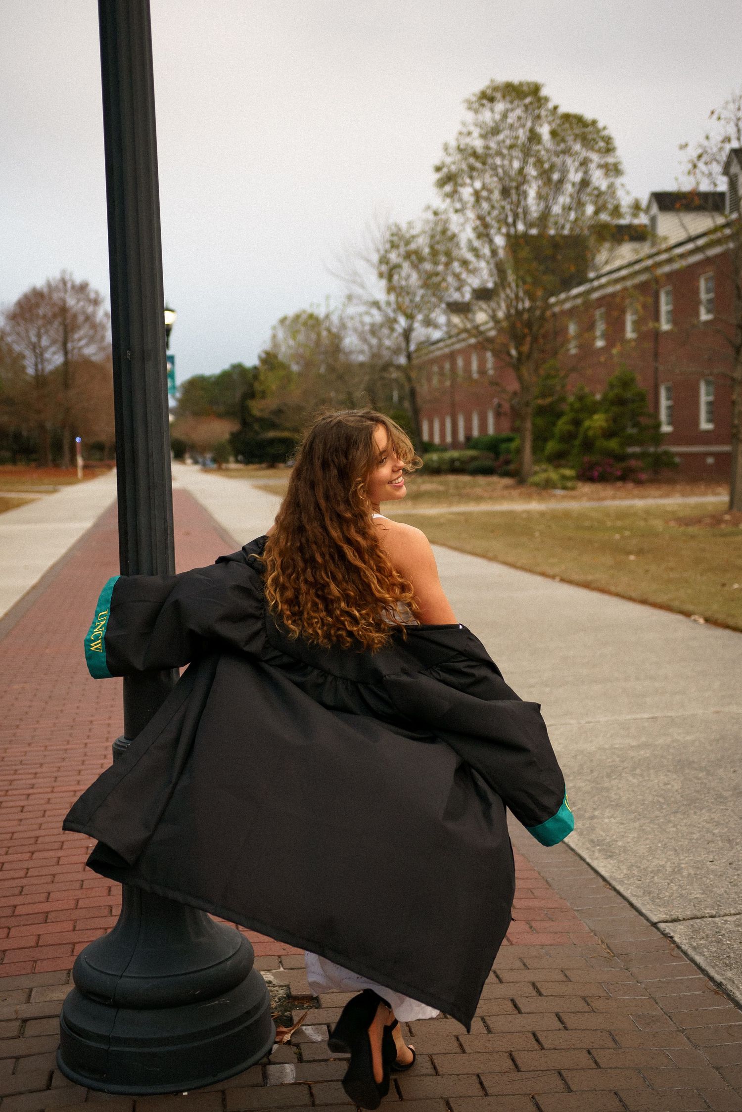 A graduation photoshoot showing a person in a black gown sitting on brick pavement near campus buildings.