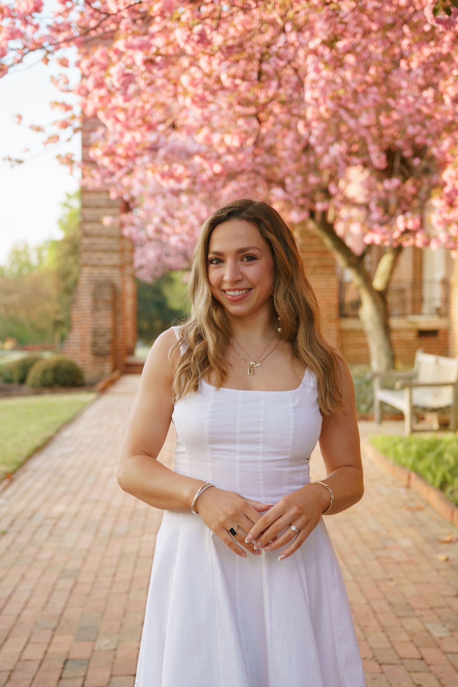 Young woman in white sundress smiling outdoors near pink cherry blossom tree on brick pathway.