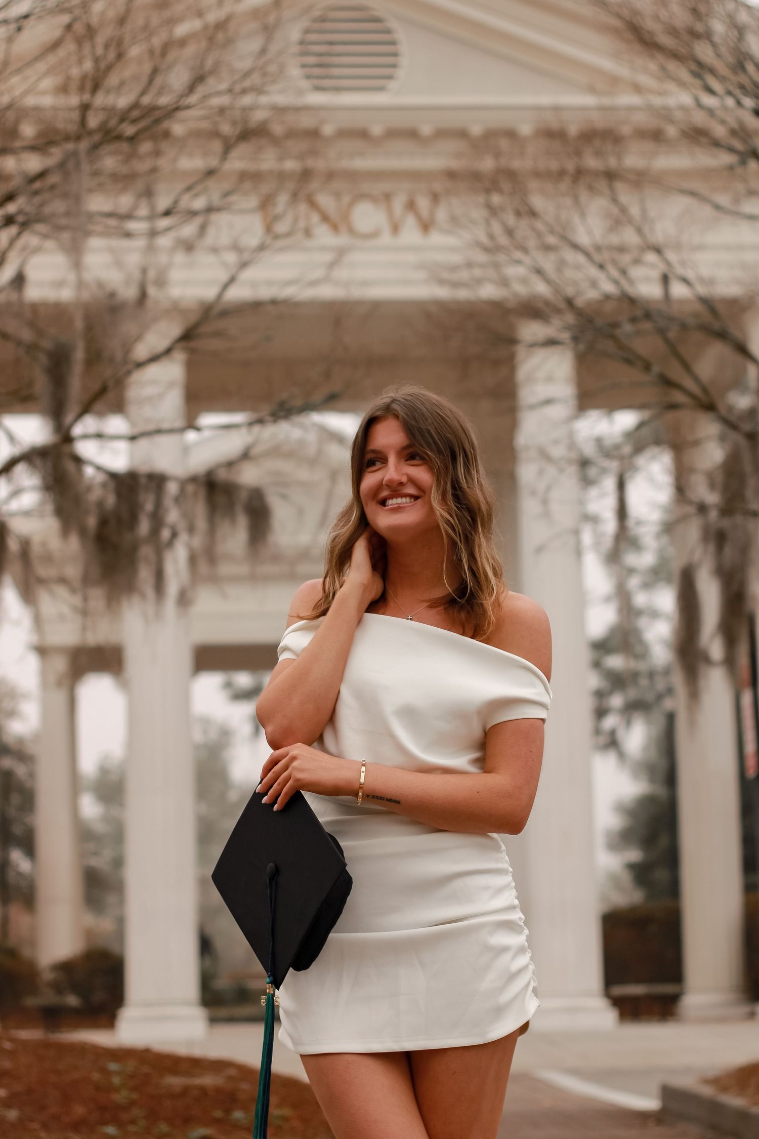 A graduate in white dress poses in front of a UNCW building with classical columns.