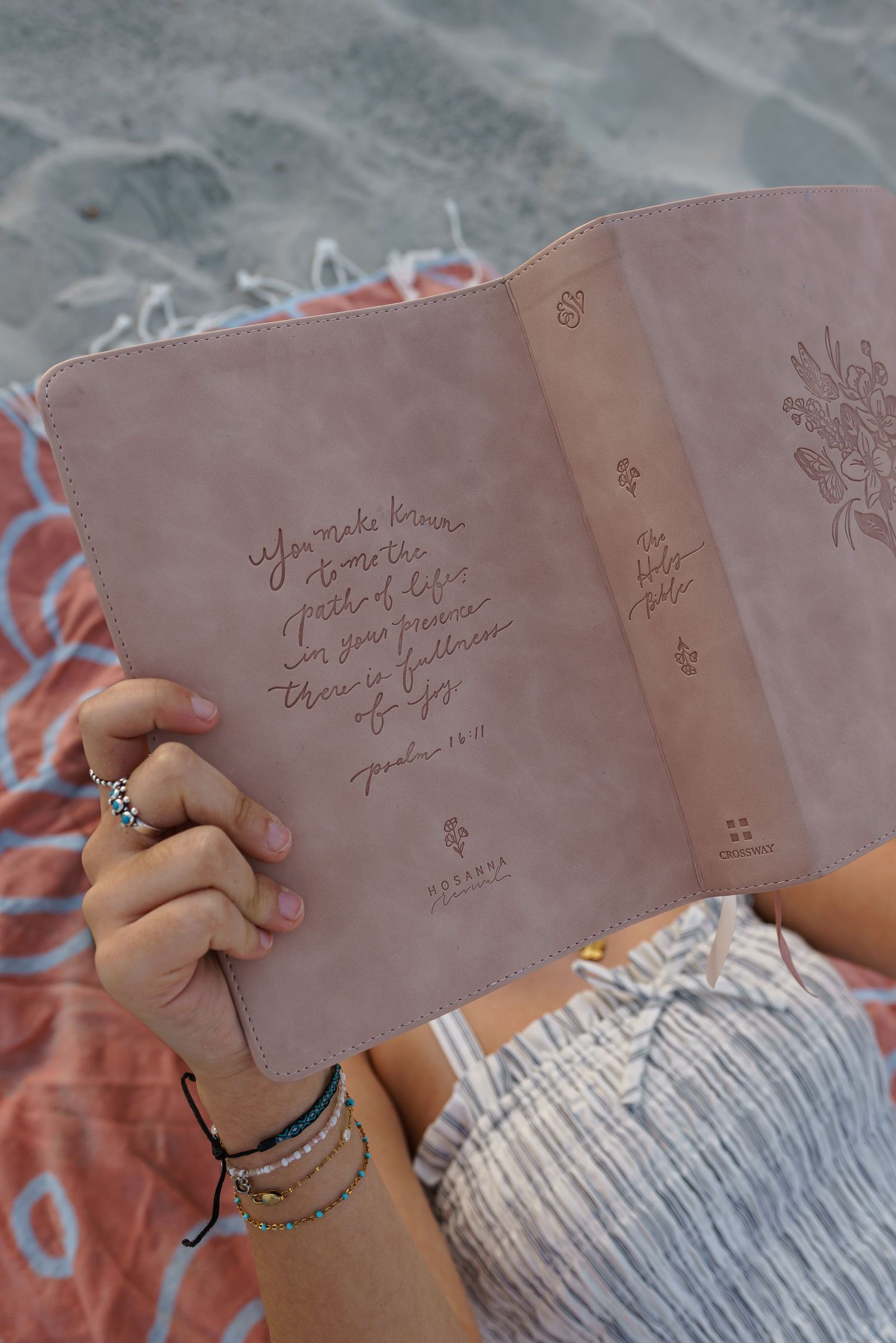 Woman reading a leather-bound Holy Bible with Psalm 16:11 inscription on sandy beach with colorful towel.