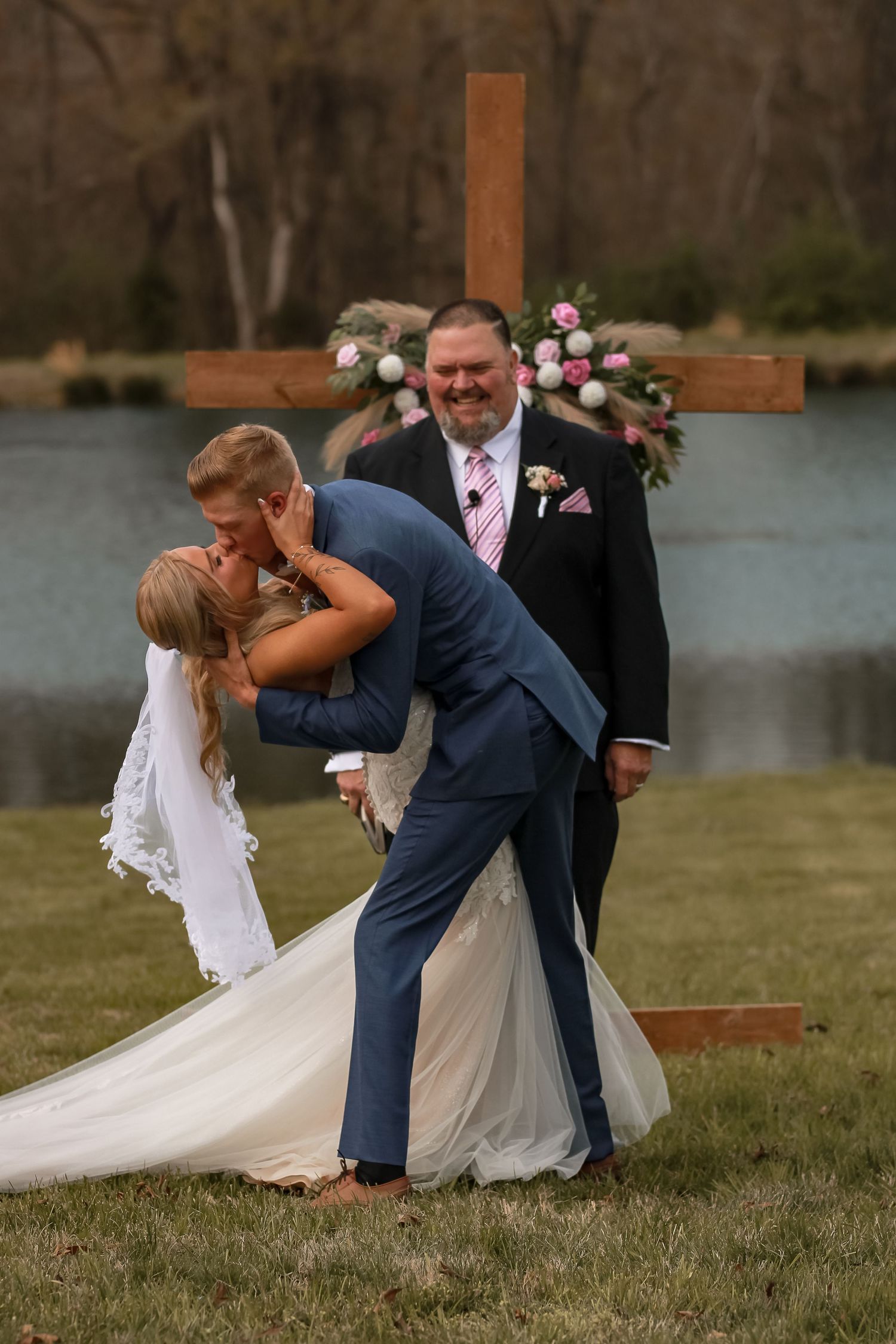 Wedding couple sharing romantic dip kiss by lakeside with wooden cross in background.