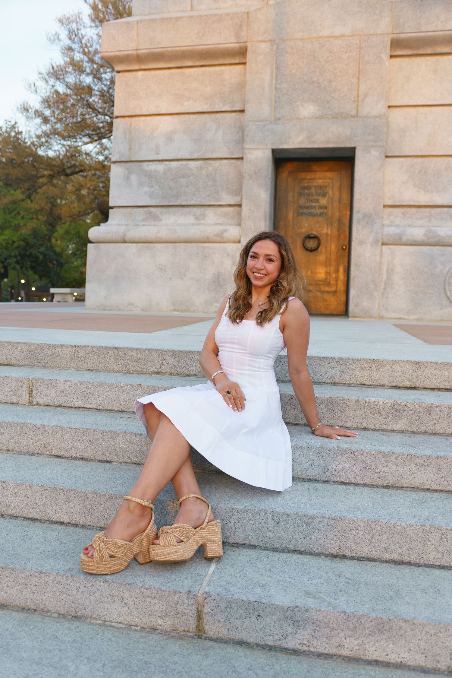 Young woman in white dress and tan platform heels sitting on stone steps outside a historic building.