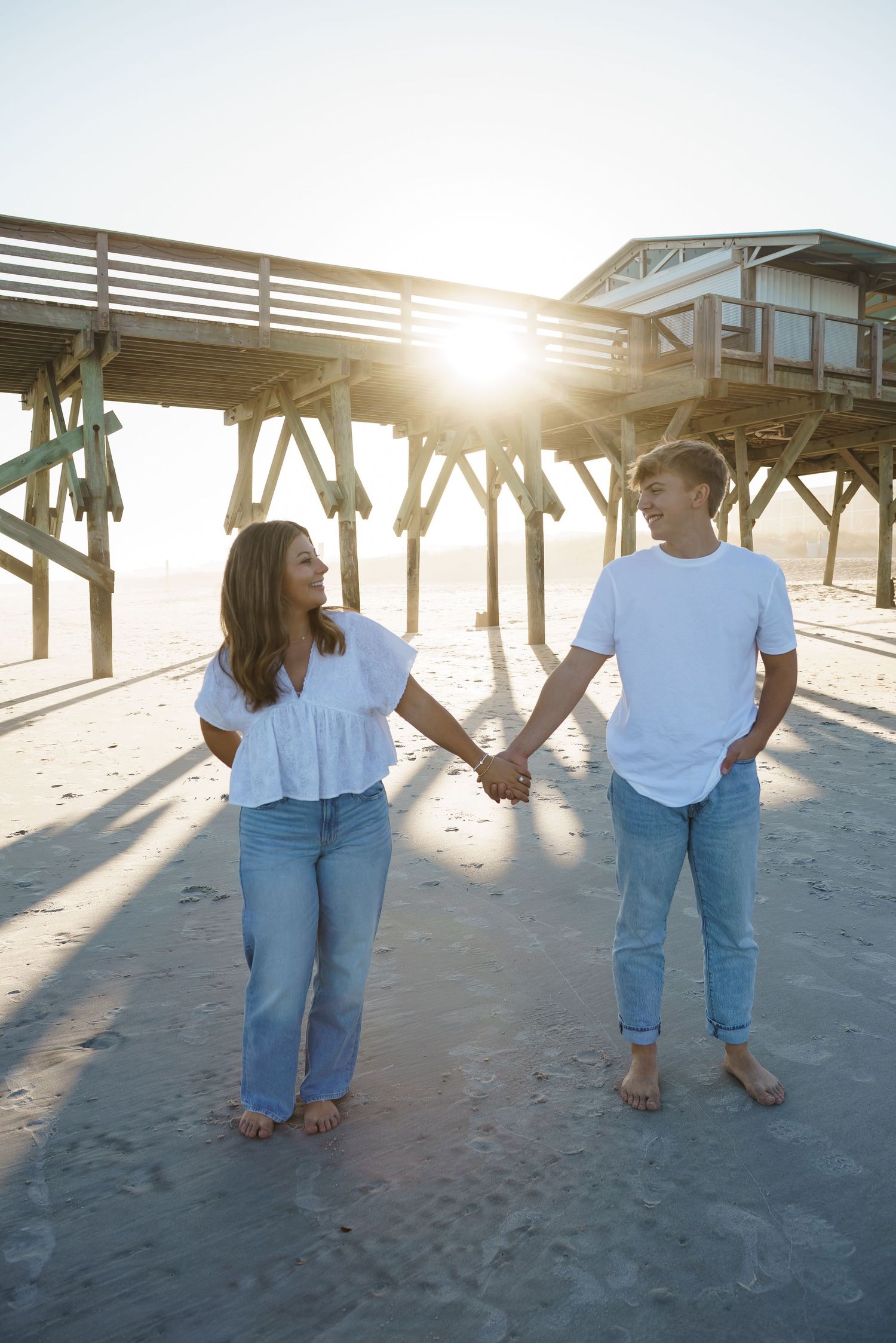 Couple walking barefoot on beach sand with pier silhouetted against setting sun.