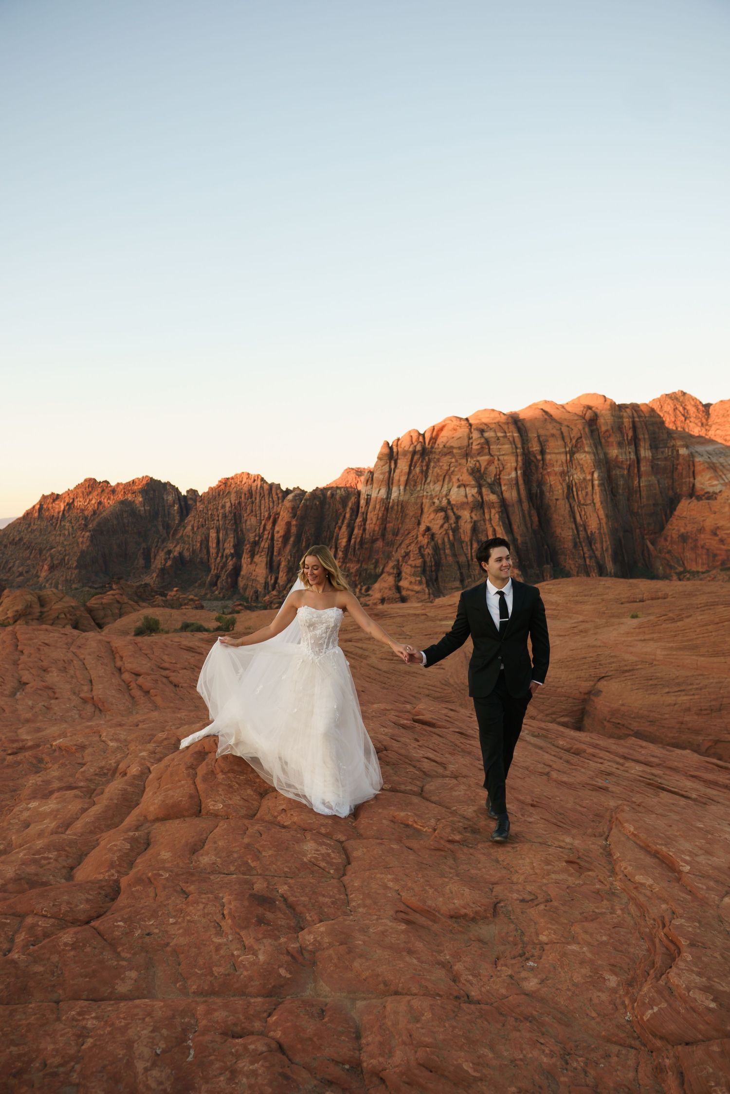 Wedding couple traverses dramatic red rock landscape during golden hour in the desert.
