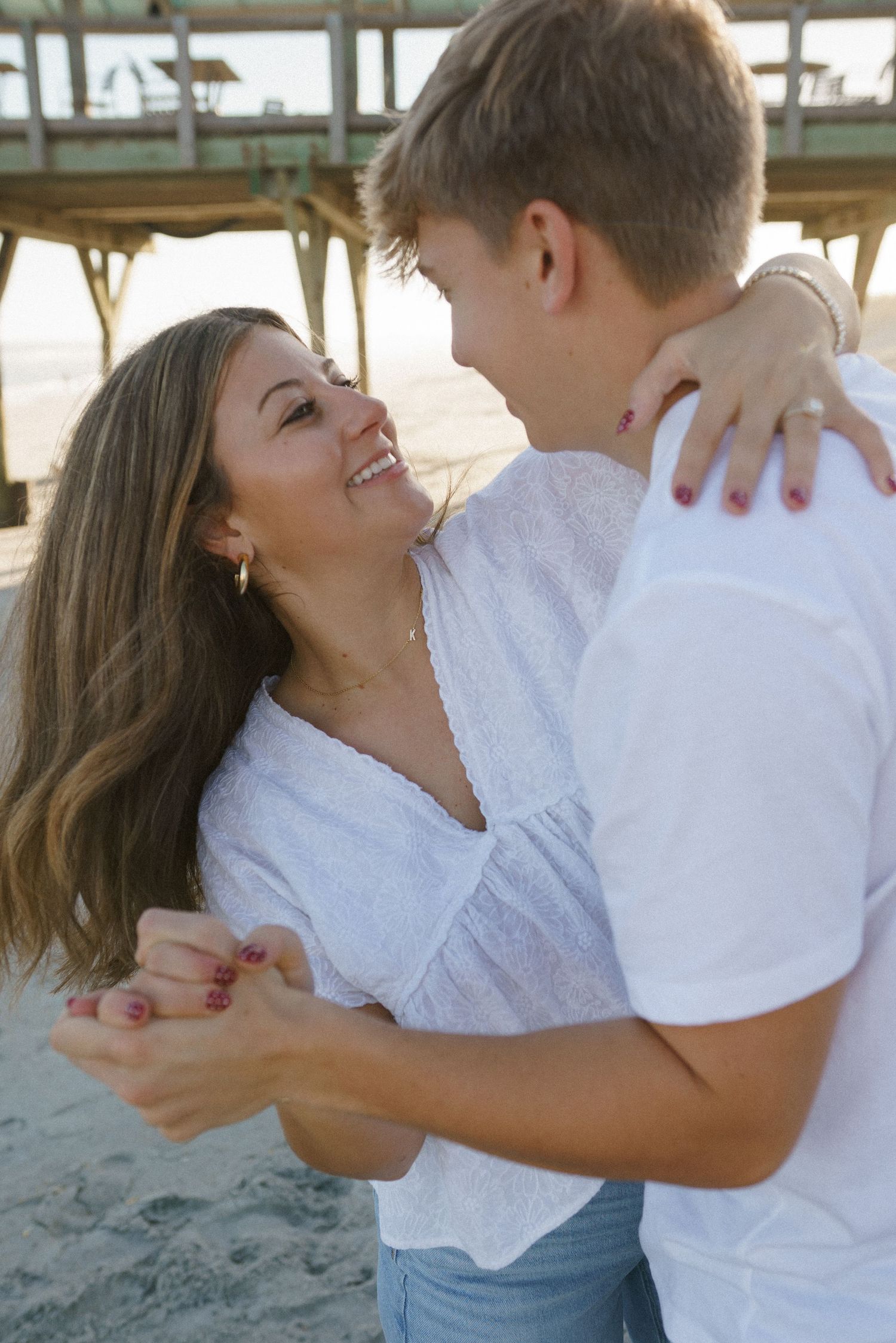 Playful couple laughing and embracing on beach with pier in background.
