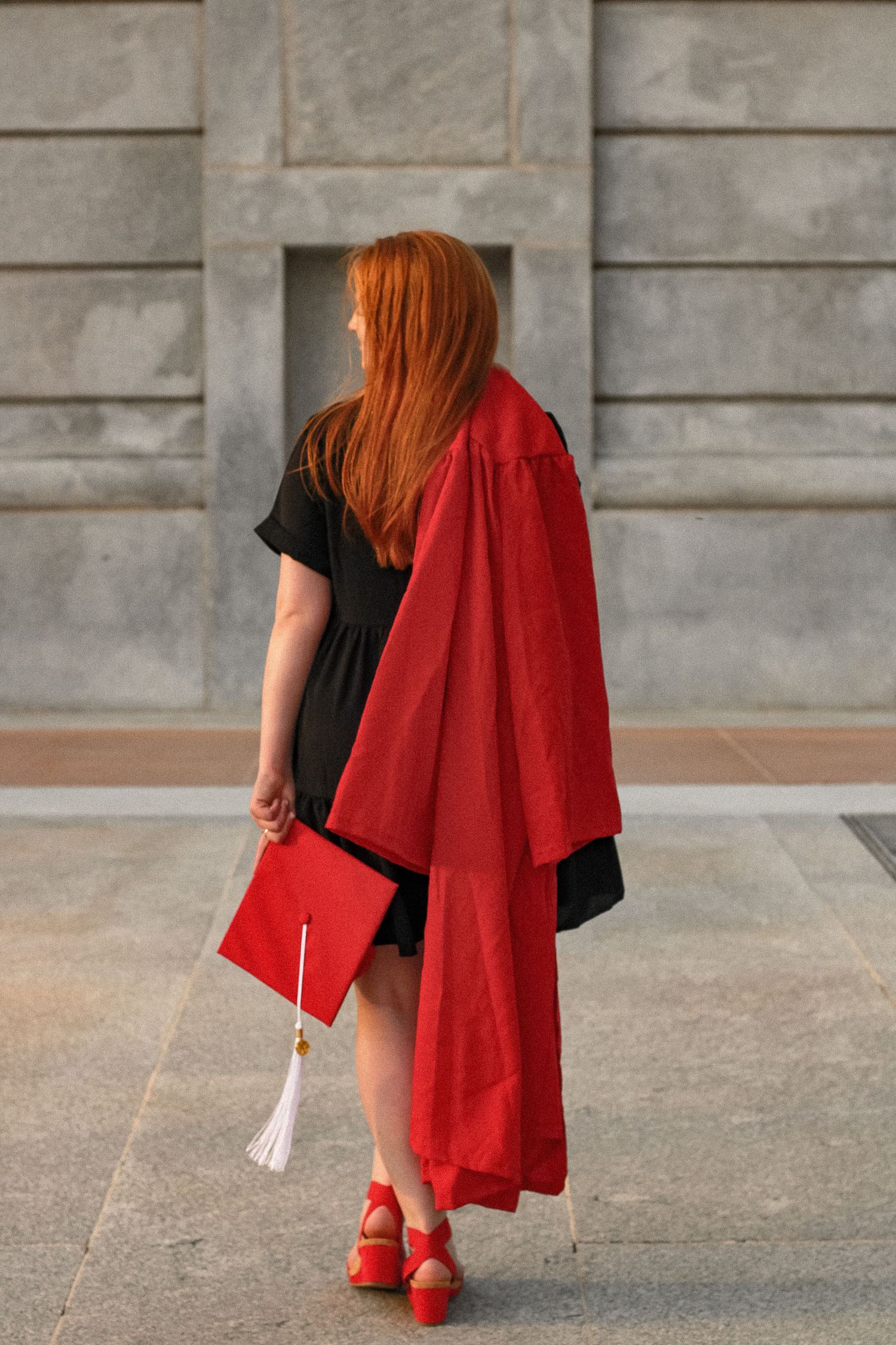 Back view of graduate holding red diploma cover wearing black dress and red graduation gown.