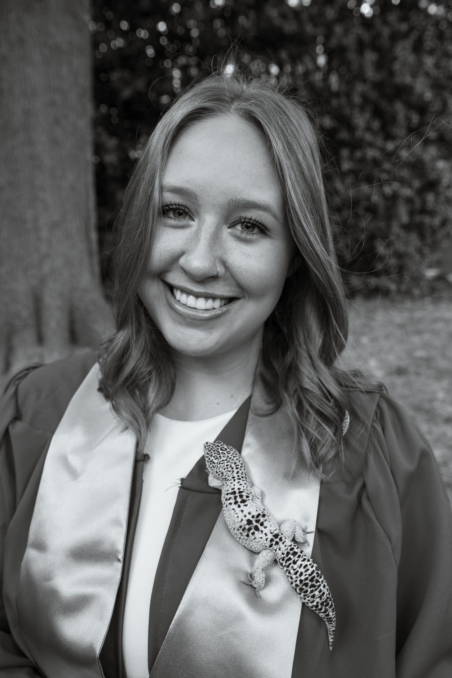 Smiling young woman in graduation gown with a leopard gecko on her chest, black and white portrait outdoors.
