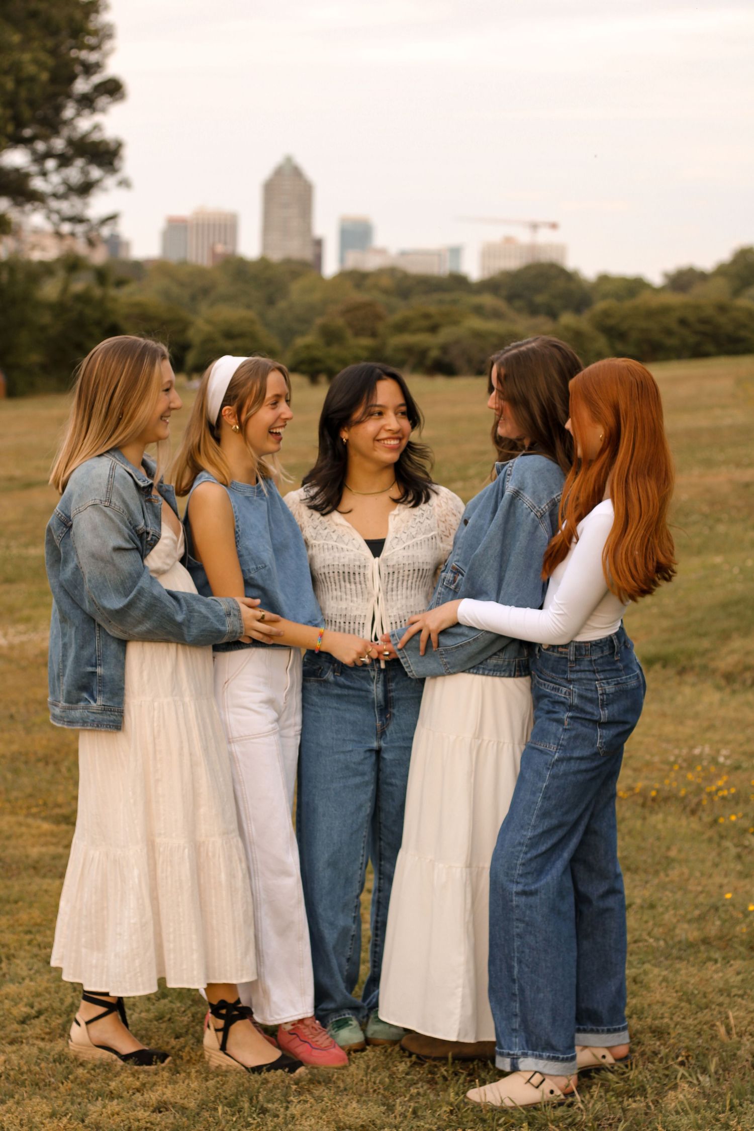 Five friends in denim and white dresses pose together with city skyline in background.