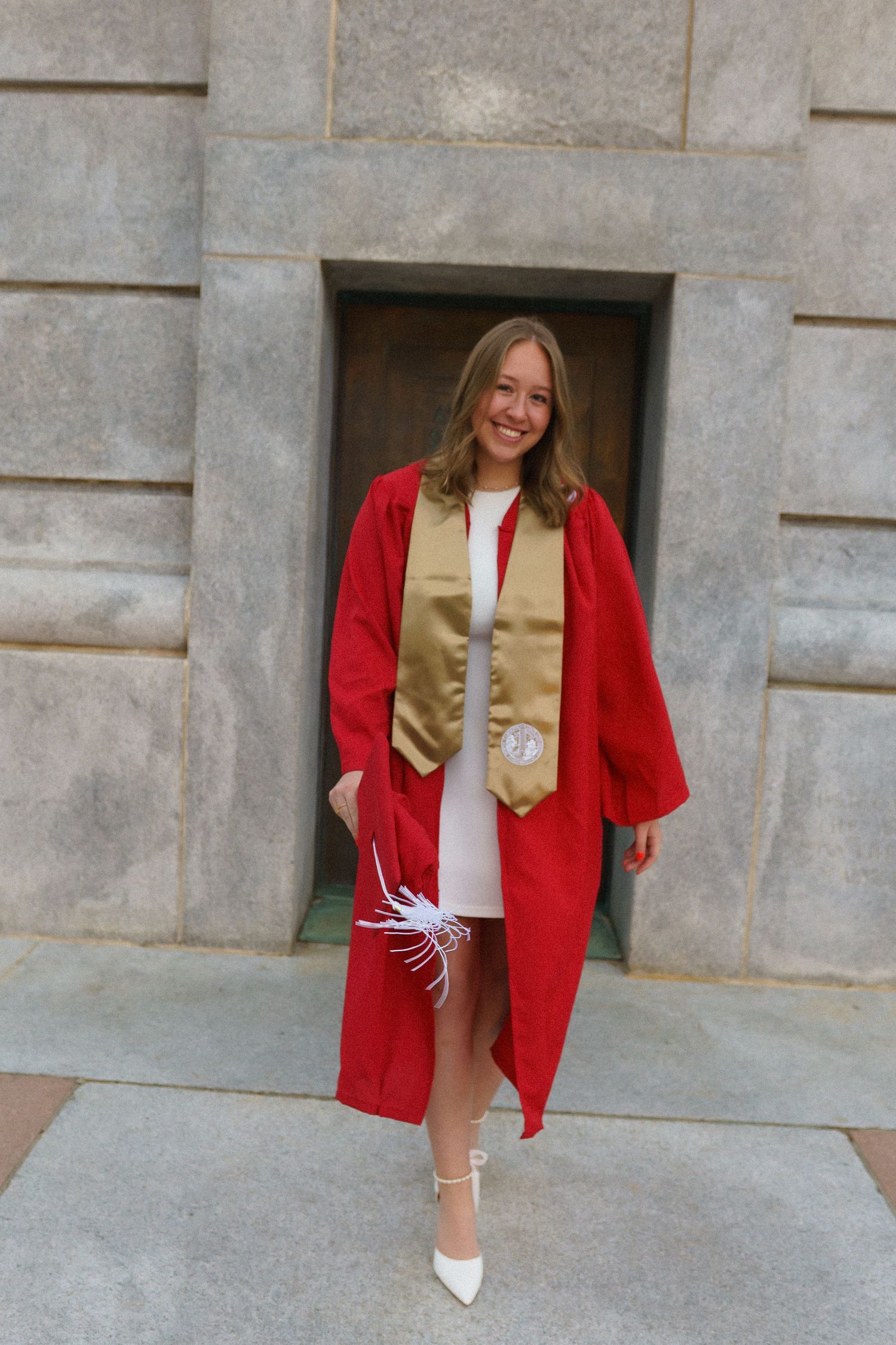 Smiling graduate in red cap and gown with gold stole holds white flowers outside stone building.