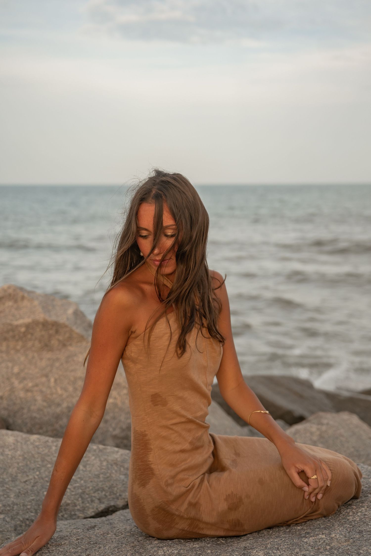 Person in brown dress sitting on rocky beach beside ocean waves.