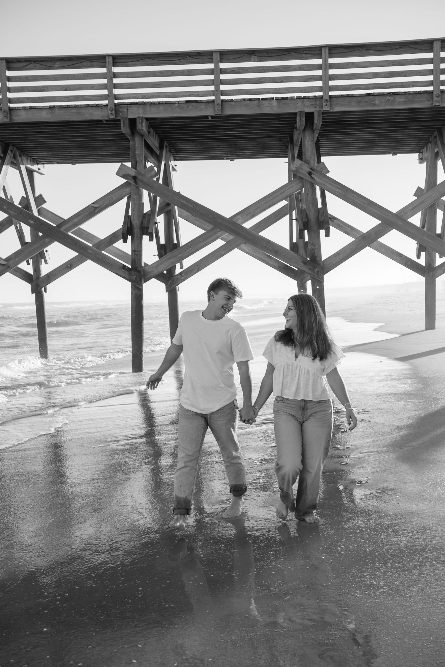Couple walking under a wooden pier with waves splashing around their feet.