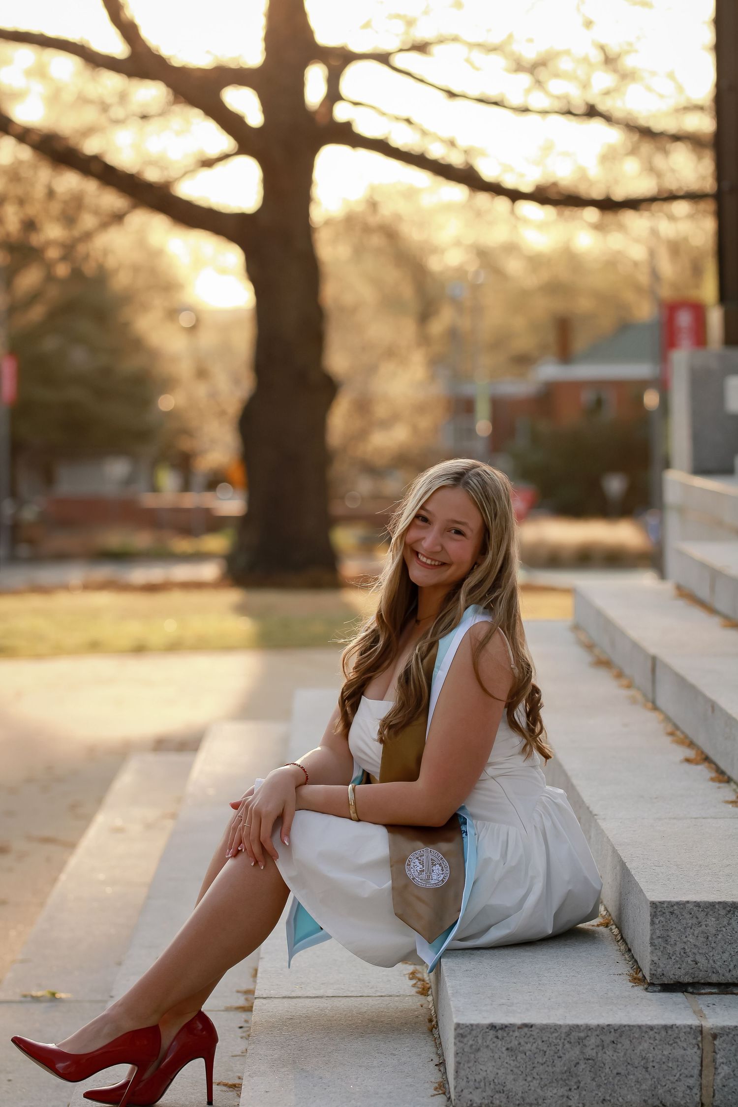 Graduate in white dress with red heels posing on concrete steps at sunset.