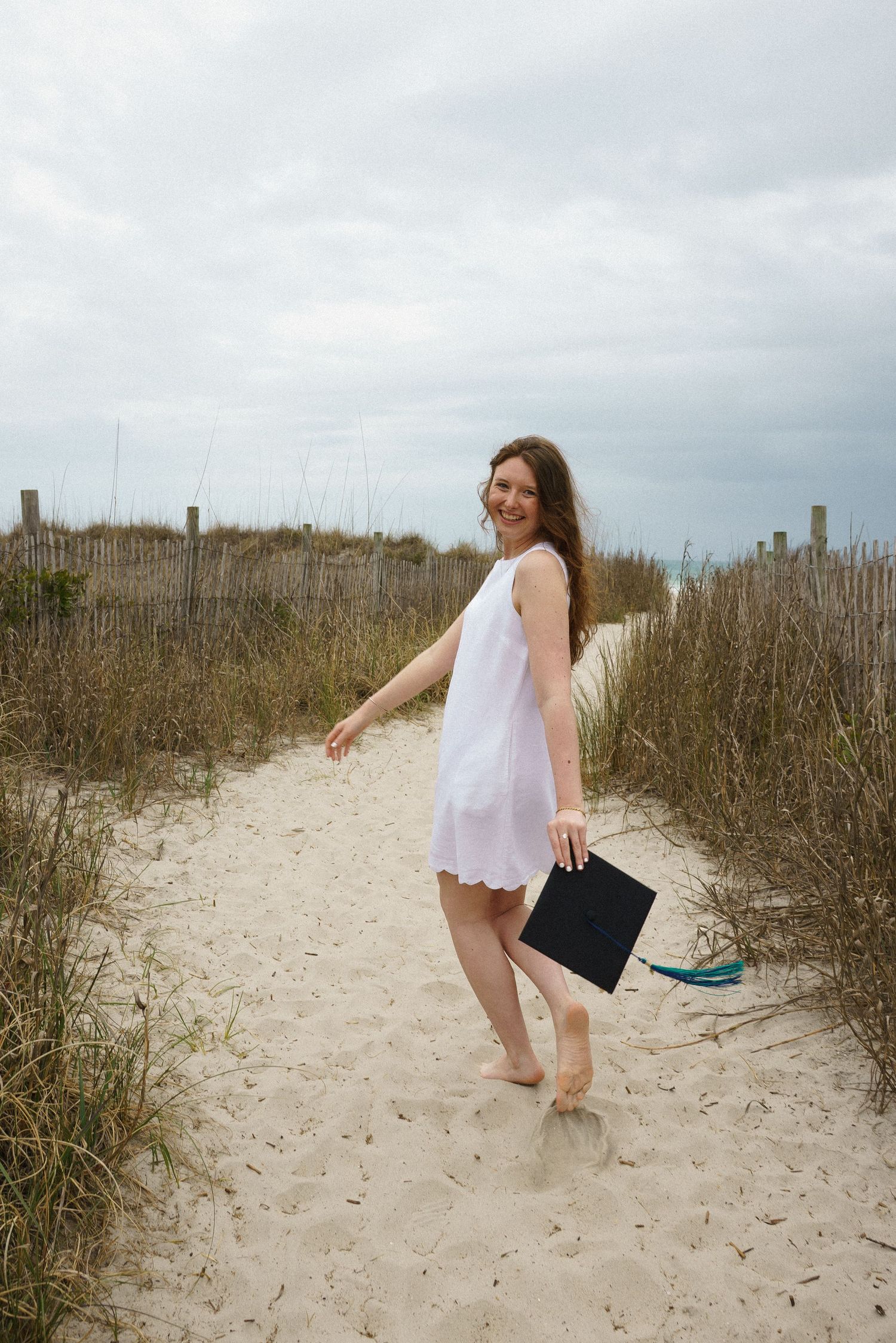 Woman in white dress holding graduation cap walks barefoot on sandy beach path with dunes and cloudy sky.