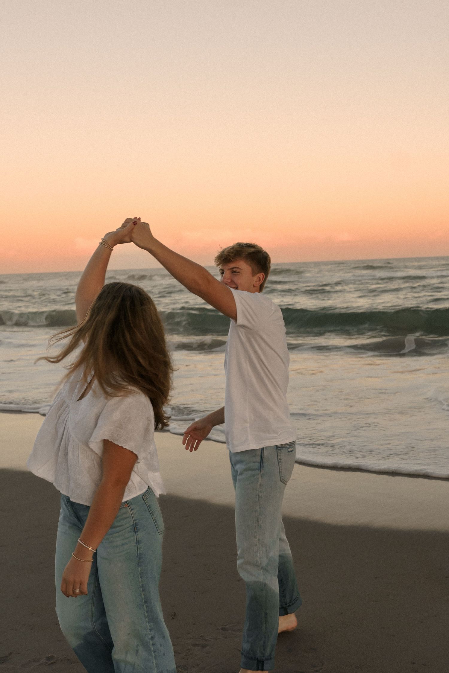 A couple dancing together on the beach at sunset in casual white shirts and jeans.