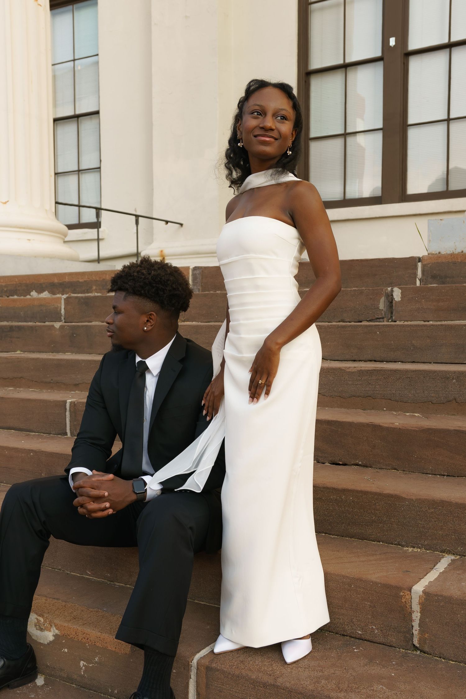Elegant couple posing on stone steps, woman in white halter gown, man in black suit, formal occasion.