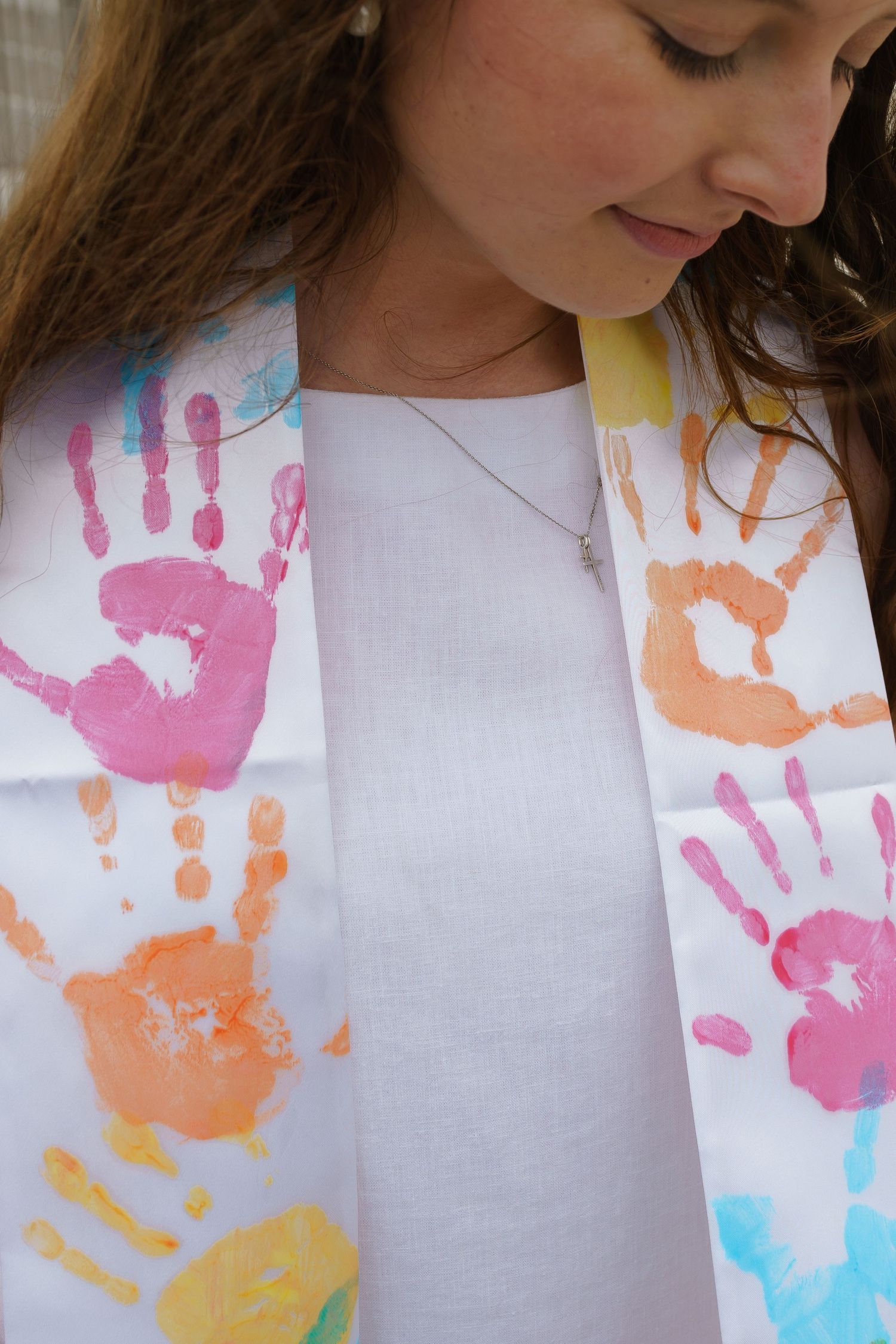 Young woman wearing a white graduation stole decorated with colorful children's handprints in pink, orange, and blue.
